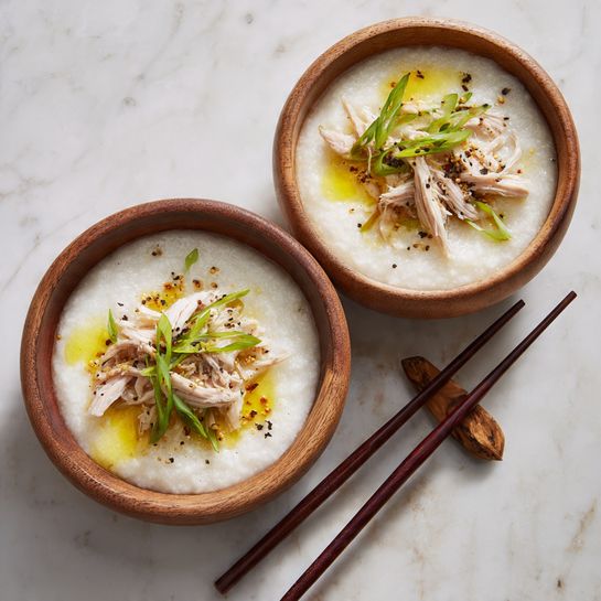 Two wooden bowls hold a smooth, white rice porridge base with a thick, soft texture. On top of the porridge in the foreground bowl are thin, light pinkish strips of shredded chicken, small bright green slices of scallions scattered around, a small pool of yellow oil, and a light sprinkle of black pepper. The bowls rest on a white marbled surface and a pair of dark wooden chopsticks are placed nearby. The photo taken with an iphone --ar 4:5 --v 7