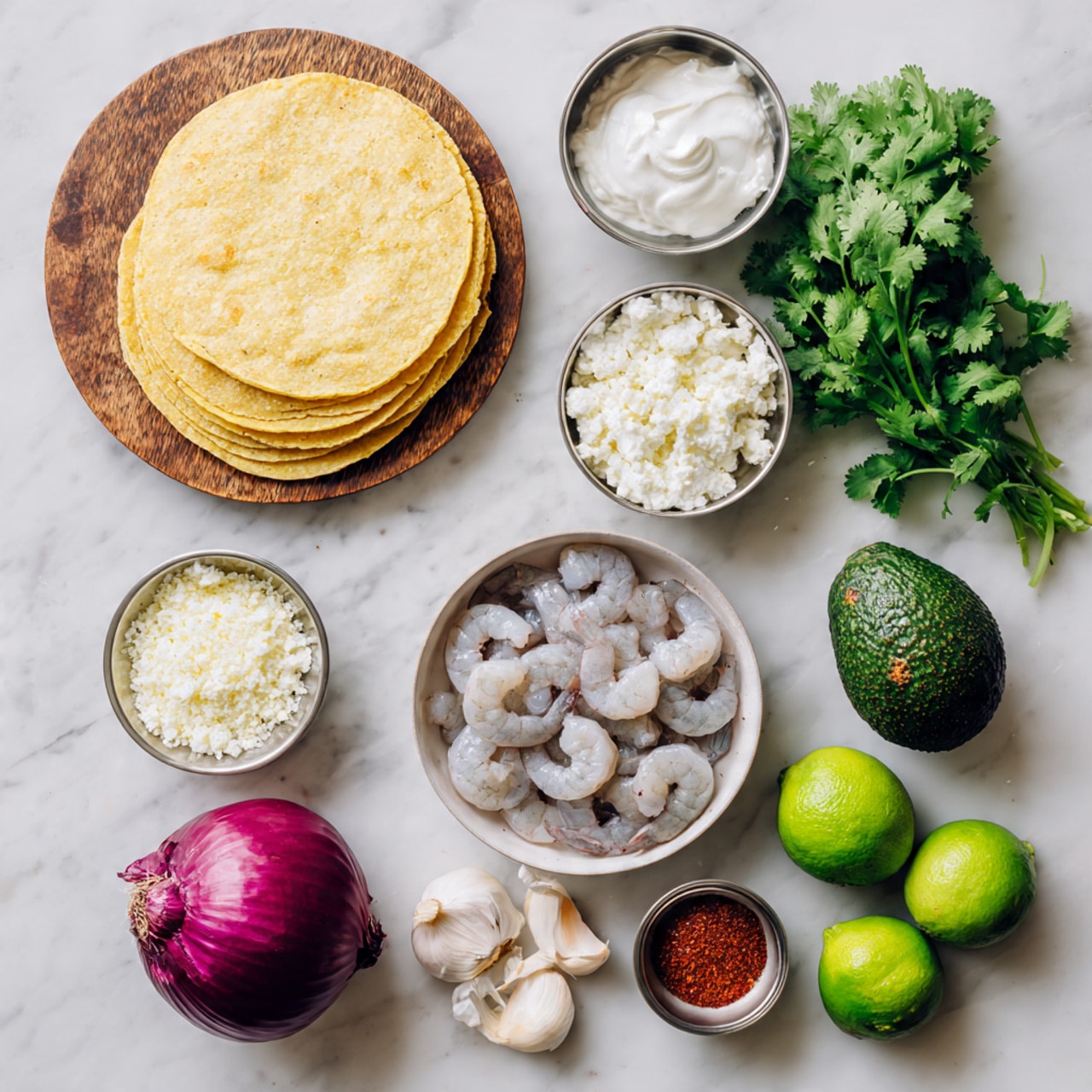A flat stack of 8 yellow corn tortillas sits on a round white plate on the left side, with a bunch of fresh green cilantro above it. To the right of the tortillas are three small metal bowls arranged vertically: the top bowl holds white sour cream with a smooth texture, the middle bowl contains crumbly white cheese, and the bottom bowl has mixed spices including black pepper, salt, and red powder. To the right of these bowls is a white bowl filled with light pink raw shrimp. Beside the shrimp, a whole dark green avocado with rough skin is placed. At the bottom left corner, half a purple onion and two green limes are arranged closely. On the top right corner, half a white onion with light orange skin edges and two garlic cloves are laid out. The whole scene is set on a white marbled textured surface photo taken with an iphone --ar 4:5 --v 7