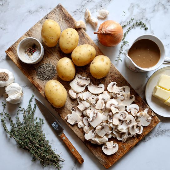 The image shows a wooden cutting board placed on a white marbled surface, filled with fresh sliced white mushrooms at the bottom, with their white caps and light brown gills clearly visible. Toward the top of the board, five whole yellow potatoes are arranged with their smooth skin. A single brown onion sits to the left side. Near the onion, there are a few garlic cloves in their purplish skin. A small beige bowl with mixed coarse salt, black pepper, and dried herbs is on the top left corner of the board. A small knife with a wooden handle rests on the board’s bottom left edge. Around the board on the white marbled surface, there is a glass measuring cup filled with brown liquid broth on the right top corner, next to a white bowl with a white creamy sauce, and a small white plate holding two pieces of butter. Fresh sprigs of thyme and rosemary are scattered around the board. Photo taken with an iphone --ar 4:5 --v 7