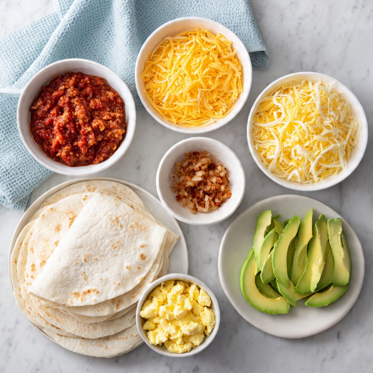 The image shows several white dishes arranged on a white marbled surface, each containing ingredients for making wraps. In the top left, there is a white bowl with a chunky red salsa. To its right, a white bowl holds shredded bright yellow cheese. Next to the cheese, a small white bowl contains small chopped pieces of a brown cooked ingredient. Below these, a stack of soft white flour tortillas is placed on the surface. At the bottom left, a white bowl is filled with scrambled eggs that are light yellow and slightly fluffy. To the bottom right, a white plate holds neatly sliced green avocado pieces. A folded light blue cloth is seen in the top left corner of the image. photo taken with an iphone --ar 4:5 --v 7