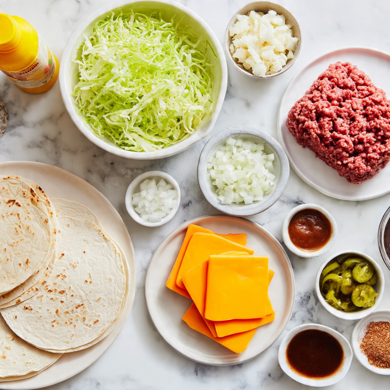 The image shows a flat lay of ingredients on a white marbled surface. There are four soft tortillas stacked on a white plate at the bottom left. Above the tortillas, a white bowl is filled with shredded green lettuce. To the right of the lettuce are several small white bowls: diced white onions, finely chopped pickles, a mix of garlic powder and onion flakes, a deep red sauce, creamy white mayonnaise, and reddish paprika powder. At the upper right, a white plate holds raw ground beef with a textured, tightly packed appearance. On the lower right, several bright yellow slices of cheese are stacked in plastic wrap. A bright yellow mustard bottle labeled
