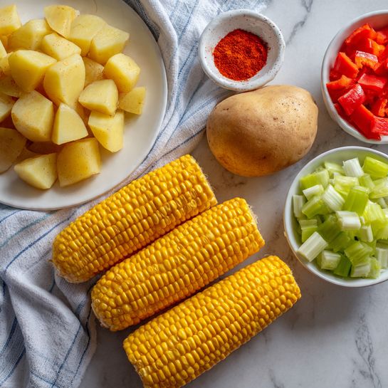 The image shows a white marbled surface covered partly by a white and blue striped cloth. On the cloth, there are four ears of yellow corn with kernels tightly packed, lying side by side in the bottom left part. To the right of the corn is a whole light brown potato with some marks on its skin. Near the top left, there is a white plate with diced pieces of yellow potato with skin bits on some pieces. Above the plate, there is a small white dish holding a pile of red paprika powder. To the right of the plate, two small white bowls rest; one contains diced light green celery pieces and the other holds diced bright red bell pepper pieces, with the bowl of peppers placed slightly lower and partially on the cloth. photo taken with an iphone --ar 4:5 --v 7