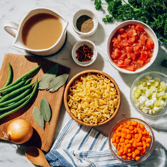 A top view of various raw ingredients arranged on a white marbled surface, including a wooden cutting board at the bottom left with a whole yellow onion, fresh green beans, two dried bay leaves, and three garlic cloves; above it are small bowls with dried herbs and red pepper flakes. To the right on the cutting board, a large white bowl filled with bright red diced tomatoes with juice, resting partly on a white cloth with blue stripes. Centered is a wooden bowl filled with small, uncooked yellow pasta tubes and a wooden spoon. Surrounding this are clear glass bowls with white beans and chopped celery, a white bowl with sliced orange carrots, and a clear pitcher of light brown broth. Fresh parsley is placed at the top left. The scene is brightly lit and organized with a sense of preparation. photo taken with an iphone --ar 4:5 --v 7