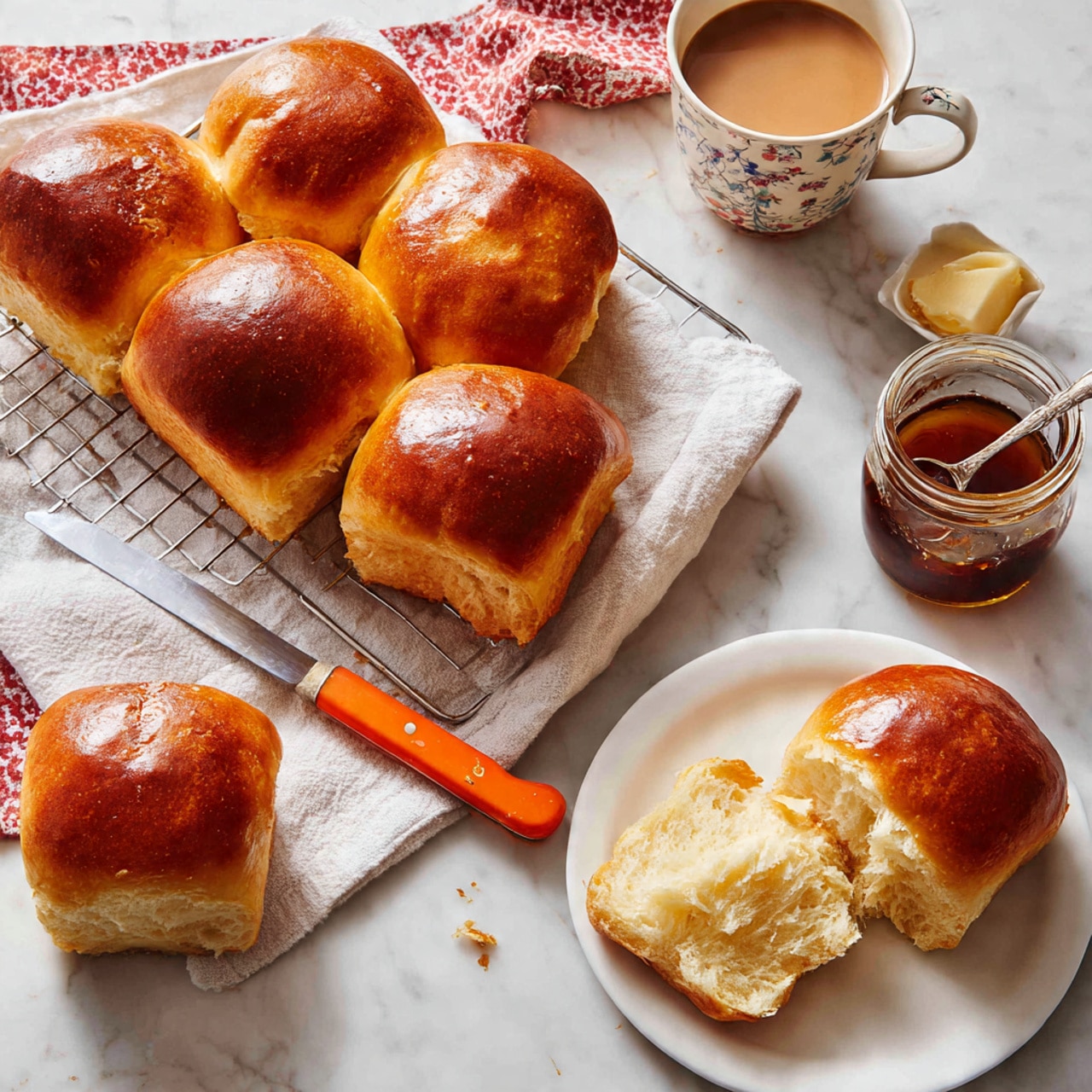 The image shows a group of seven golden brown bread rolls with a smooth shiny crust, arranged on a white wire cooling rack with a white cloth underneath, all placed on a white marbled textured surface. One roll is split open on a white plate at the bottom right, showing its soft, airy inside with a light cream color. A knife with an orange handle lies across the plate. Nearby, a white cup with a floral pattern is filled with light brown coffee. There is a small glass jar of honey with a wooden top to the right of the plate. To the left, another whole roll sits on a red and white patterned cloth next to a small spoon inside a clear jar of dark jam. A butter knife with an orange handle and a small piece of butter rests near the rolls. The scene has a cozy homemade feel with soft natural light. Photo taken with an iphone --ar 4:5 --v 7