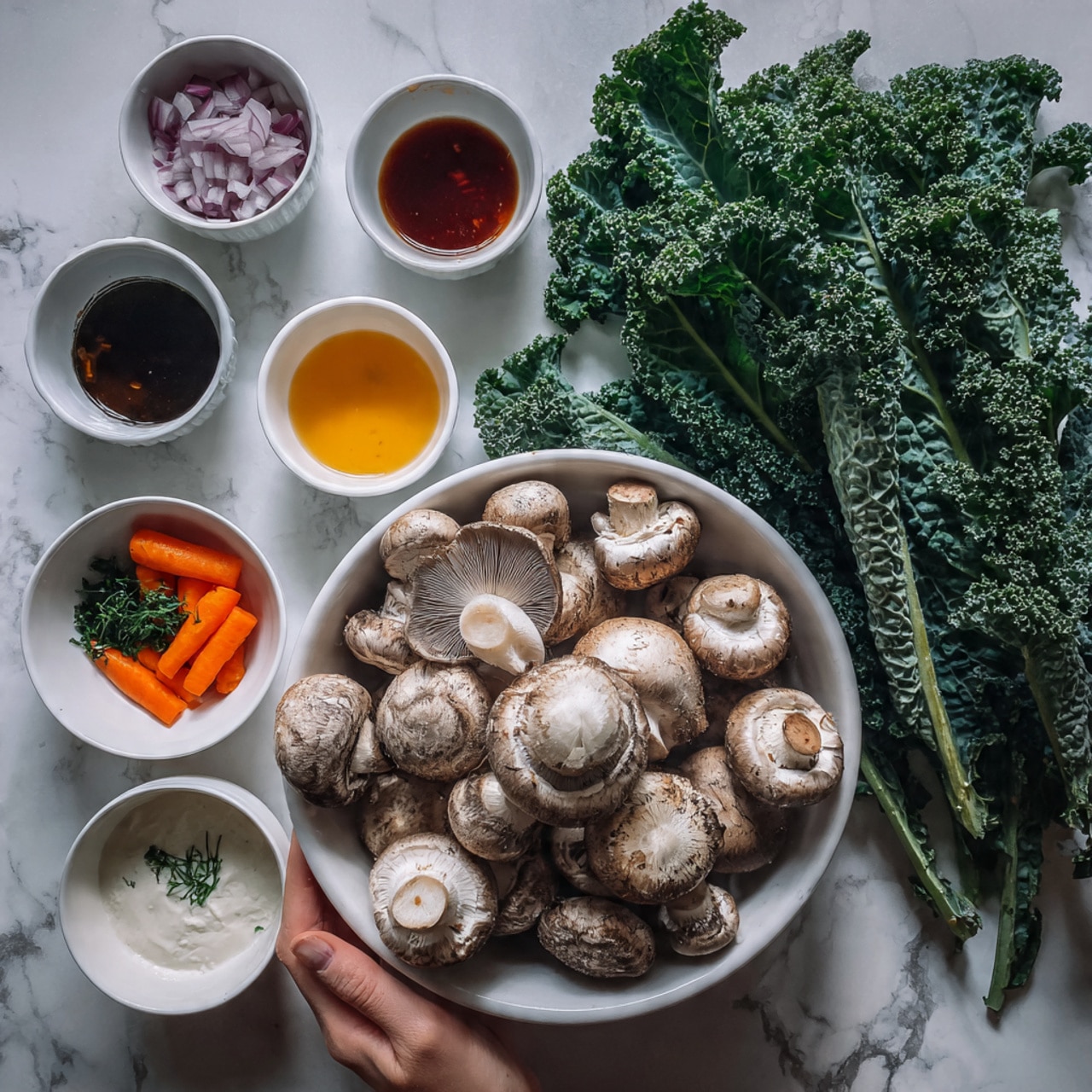 The image shows a white bowl filled with fresh whole mushrooms placed on a white marbled surface. Nearby, a large bunch of green kale with textured curly leaves lays spread out. On the side, there are small white bowls containing various ingredients: one with chopped red onions, one with a golden yellow oil, one with two orange carrots, and one with a mix of fresh white cream and dark green herbs. Small cups with colorful sauces, including a dark brown and a bright red one, are also arranged around. A woman's hand is gently touching the kale. Photo taken with an iphone --ar 4:5 --v 7