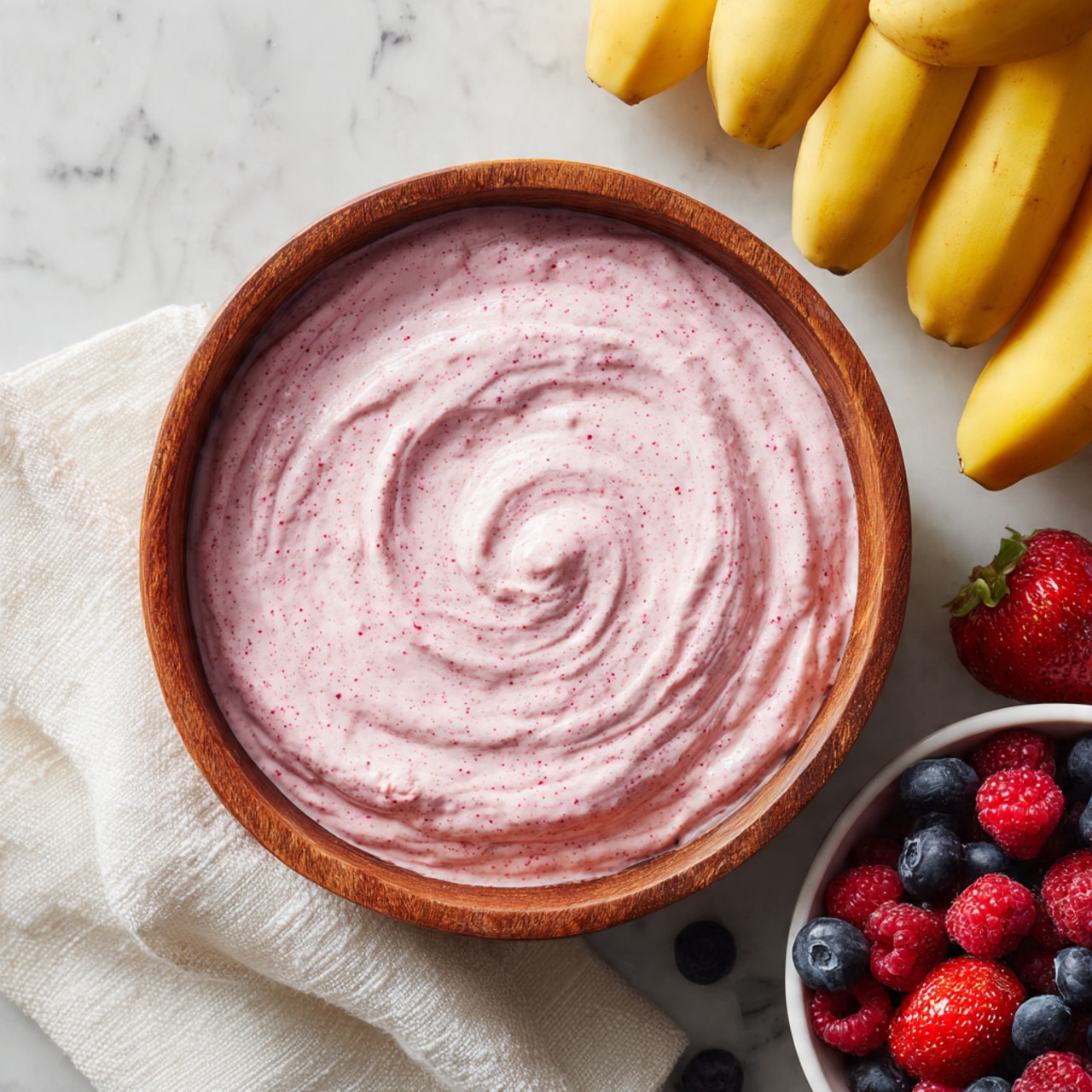 A round wooden bowl filled with smooth, light pink creamy mixture that has small red specks, swirled gently in the center; to the right, a bunch of yellow bananas on a white textured cloth; at the bottom right, a white bowl filled with fresh strawberries, blueberries, and raspberries showing bright red and deep blue colors; all items placed on a white marbled surface, photo taken with an iphone --ar 4:5 --v 7