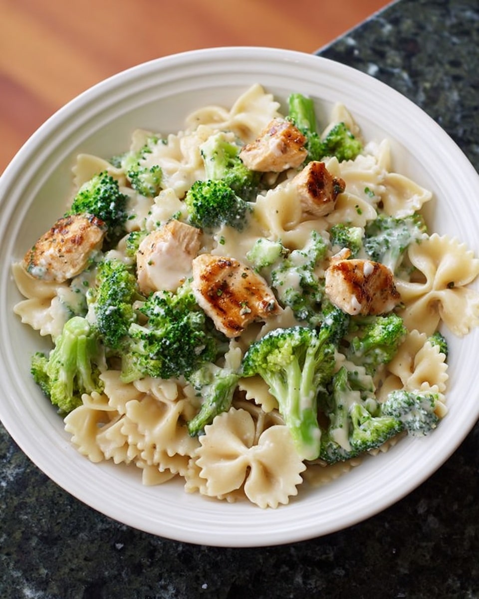 A white plate filled with farfalle pasta covered in a creamy white sauce, with vibrant green broccoli pieces mixed evenly throughout. On top of the pasta and broccoli, there are small pieces of light brown, grilled chicken. The pasta and sauce have a smooth texture, while the broccoli looks fresh and crisp. The plate is set on a white marbled surface. photo taken with an iphone --ar 4:5 --v 7