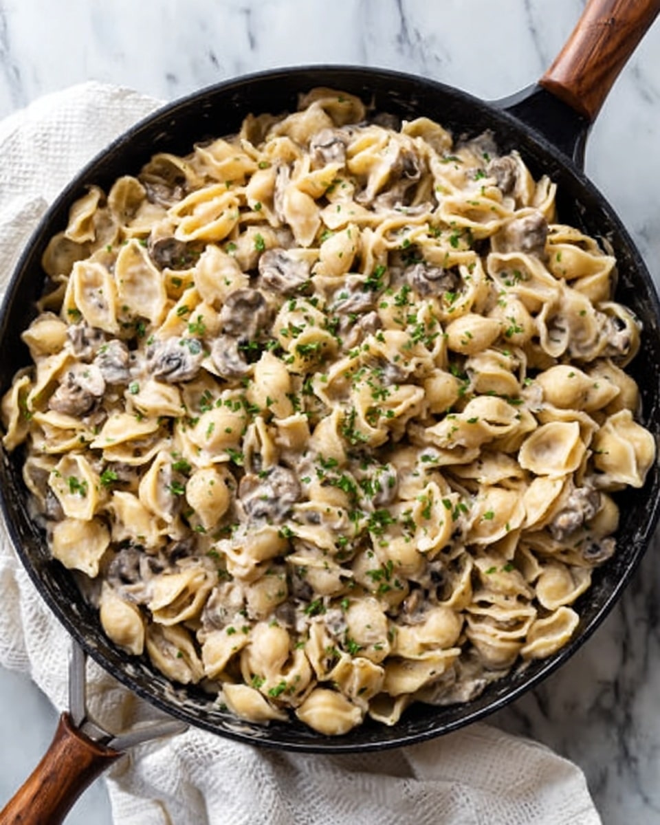 A large black pan filled with creamy pasta shells mixed with sliced mushrooms and small green herb pieces on top, creating a soft, textured look. The pasta has a light beige color with a smooth sauce coating, and the mushrooms add darker shades of brown and light gray, scattered evenly throughout. The pan sits on a white marbled surface, with a small white cloth partially visible nearby. photo taken with an iphone --ar 4:5 --v 7