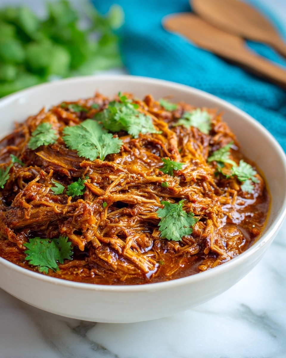 The image shows a white bowl filled with shredded meat covered in a thick brown sauce. The meat is layered densely, giving a rich and hearty look with its textured strands clearly visible. On top, a few fresh green cilantro leaves are placed as garnish, adding a splash of color. Behind the bowl, wooden utensils and a blue cloth are softly blurred, resting on a white marbled surface. The overall mood is warm and rustic, focusing closely on the detail of the cooked meat. photo taken with an iphone --ar 4:5 --v 7