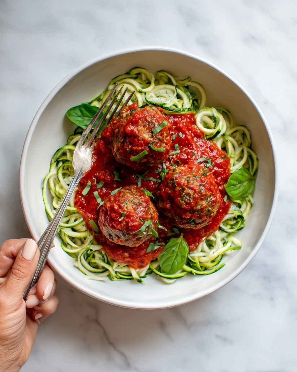 A white shallow bowl with three large meatballs covered in bright red tomato sauce centered on top of a bed of spiralized zucchini noodles that are pale green with smooth texture. Bright green basil leaves are placed around the meatballs and noodles. A silver fork with a woman’s hand holding it is visible on the bottom edge of the bowl. The bowl is set on a white marbled surface. photo taken with an iphone --ar 4:5 --v 7