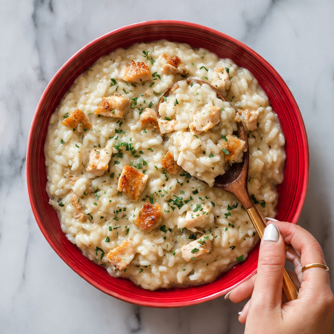 The image shows a red dish filled with creamy risotto topped with golden brown pieces of chicken. The risotto looks soft and creamy with a light beige color, and it is sprinkled with small green herbs on top. A wooden spoon is scooping some of the risotto and chicken, with a woman's hand holding the spoon gently. The background surface is a white marble texture. Photo taken with an iphone --ar 4:5 --v 7