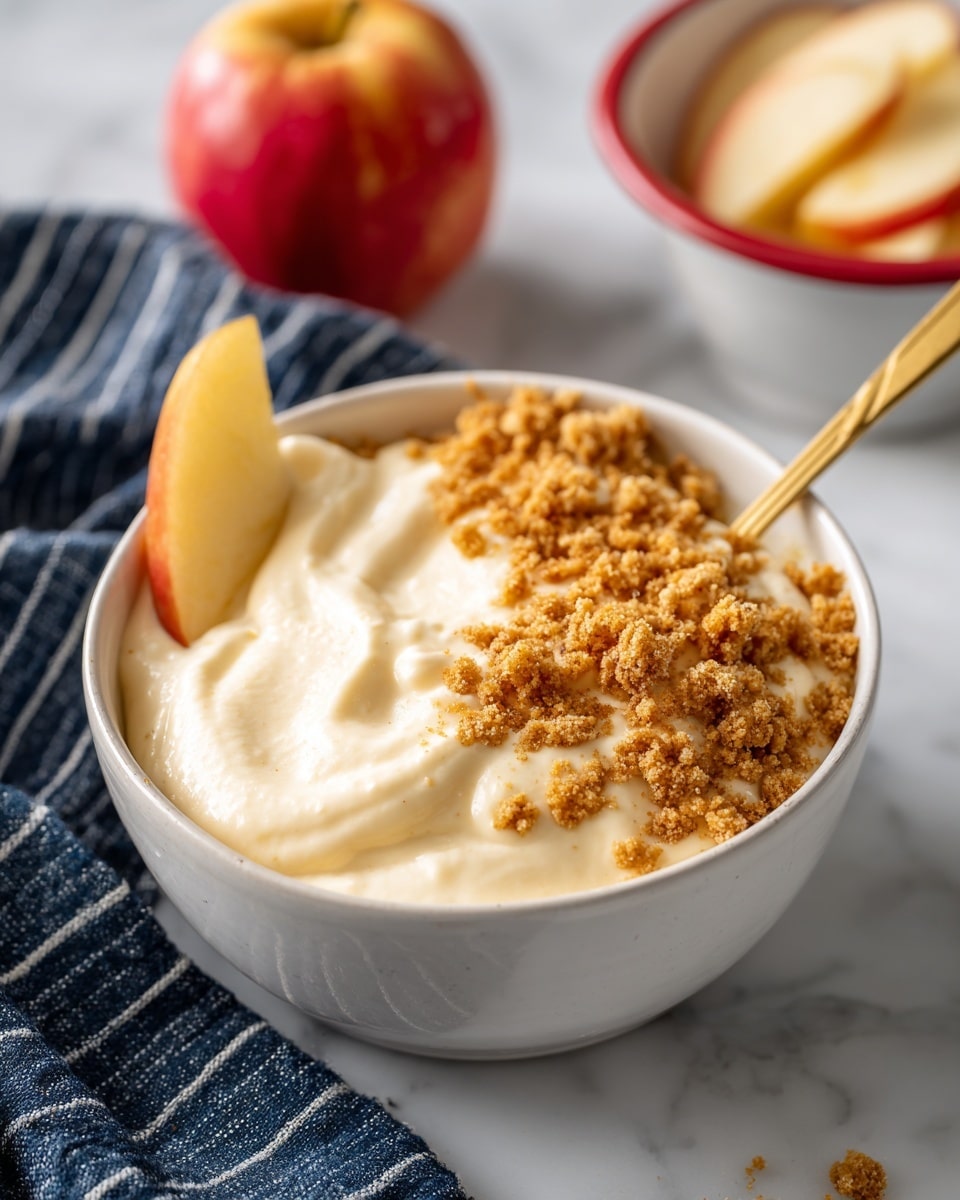 A white bowl filled with a creamy, light beige mixture that looks smooth and thick, topped with a layer of crumbled light brown crumbs on the right side and around the edges. A single pale yellow apple slice leans against the left edge inside the bowl. A gold spoon is partially submerged on the right side of the mixture. In the background, there is a white bowl with red trim holding more apple slices, and a whole red and yellow apple sits on the white marbled surface next to the bowl. A dark blue and white striped cloth is placed to the left side of the bowl. photo taken with an iphone --ar 4:5 --v 7