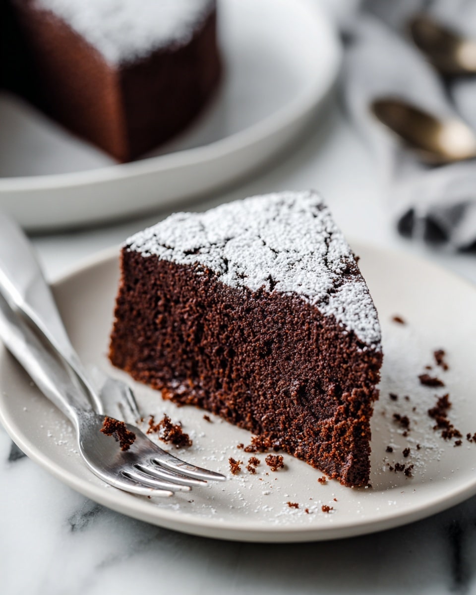 A thick, dark brown chocolate cake slice sits on a white plate with a few crumbs around it. The cake has a soft, dense texture with a slight crack on top, dusted with a light layer of white powdered sugar. Next to the cake slice are two small silver forks placed casually on the plate. The background shows a white marbled surface with blurred kitchen tools in the back. The image captures a close-up view with soft lighting highlighting the cake's rich texture. photo taken with an iphone --ar 4:5 --v 7