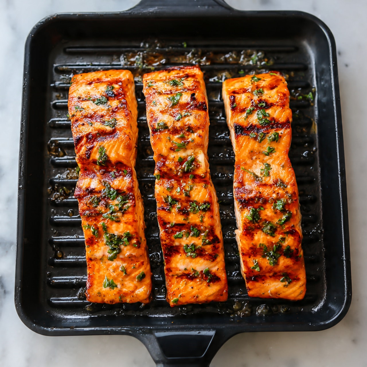 The image shows four rectangular pieces of grilled salmon placed on a black grill pan. Each piece has a bright orange color with some darker brown grill marks and a slightly crispy texture on the top surface, sprinkled with small green herb bits. The salmon looks juicy and well-cooked, giving off a shiny glaze from the oils and spices on it. The background is the black grill pan with some oil and sauce residue, and the surface under the pan is a white marbled texture. photo taken with an iphone --ar 4:5 --v 7