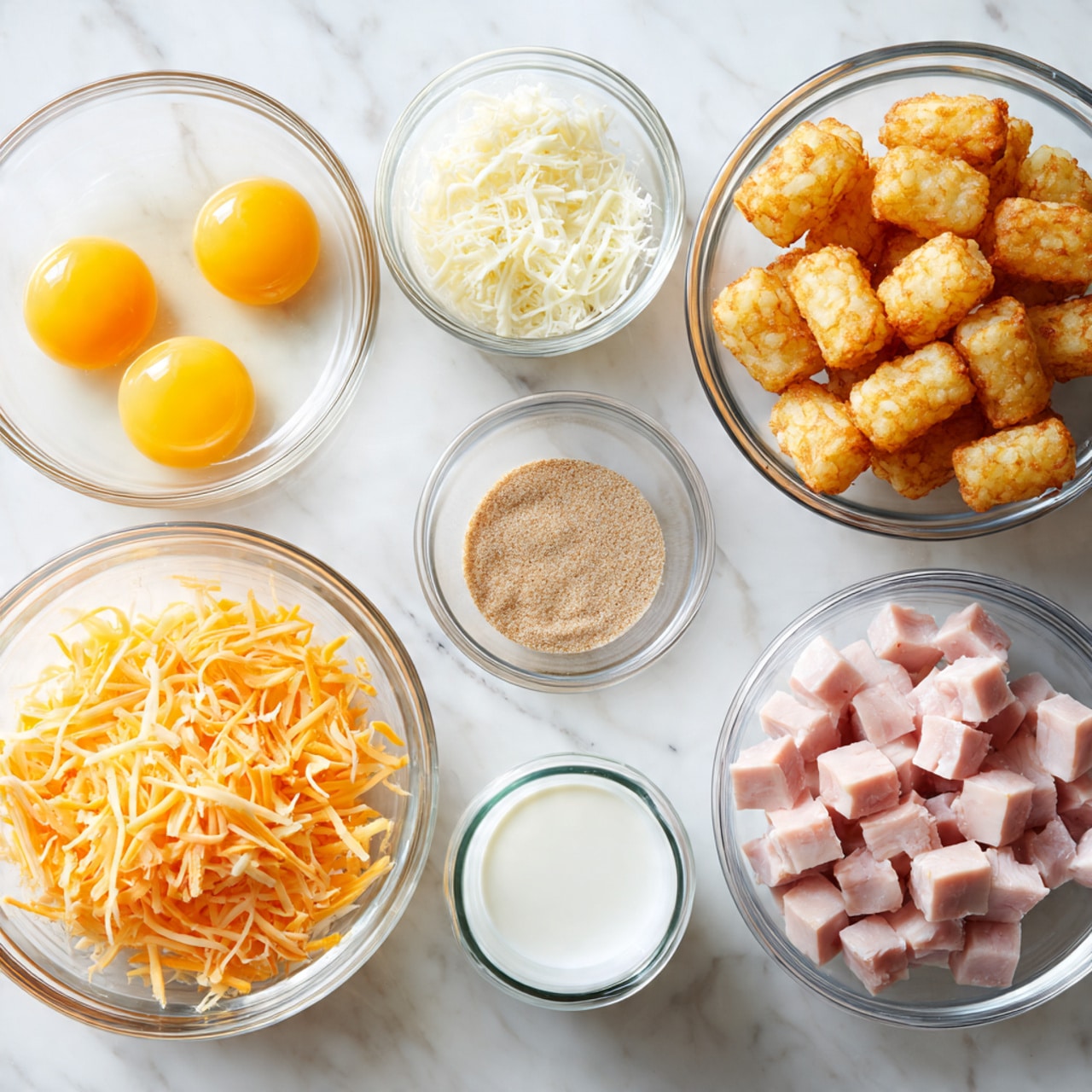The image shows six clear glass bowls arranged on a white marbled surface. One bowl contains golden brown tater tots piled up, another holds six raw yellow egg yolks with some egg white visible, and a third bowl has small cube pieces of pale pink ham. Nearby, a larger bowl is filled with shredded orange and white cheese. There is also a small bowl with a light brown seasoning powder and a small glass cup with white milk. A woman's hand is not visible in the image. The overall scene is bright and clean, emphasizing the different textures and colors of the ingredients. photo taken with an iphone --ar 4:5 --v 7
