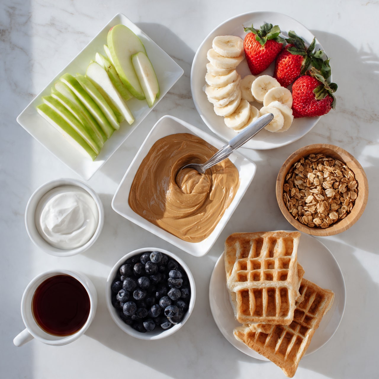The image shows a white marble surface with a white square bowl in the center filled with light brown peanut butter and a spoon inside. Around it, there is a white plate at the top left holding green apple slices and red strawberry halves with green leaves. At the top right, there is a white plate with banana slices arranged in circles. A small wooden bowl on the right side contains light brown granola. Below the peanut butter, there is a small bowl filled with dark blue blueberries, and next to it on the right, there is a white plate with two light brown toasted waffles stacked. On the left side, there is a small white square bowl with white cream and a cup with a dark red liquid. Photo taken with an iphone --ar 4:5 --v 7
