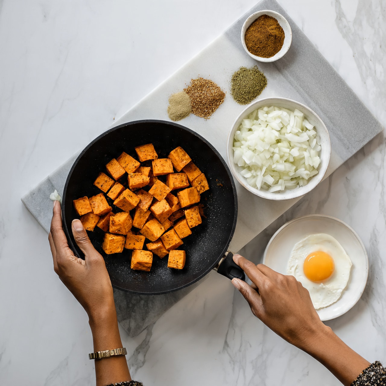 The image shows a black pan on a white marble surface. Inside the pan are small orange sweet potato cubes placed at the top left corner. Next to the sweet potatoes, on the white marble surface, is a white bowl with chopped white onions. Below the bowl with onions is a white plate holding an egg and several small piles of different spices in shades of brown, green, and beige. A woman’s hand is reaching from the left side of the image, holding onto the pan’s handle. Photo taken with an iphone --ar 4:5 --v 7