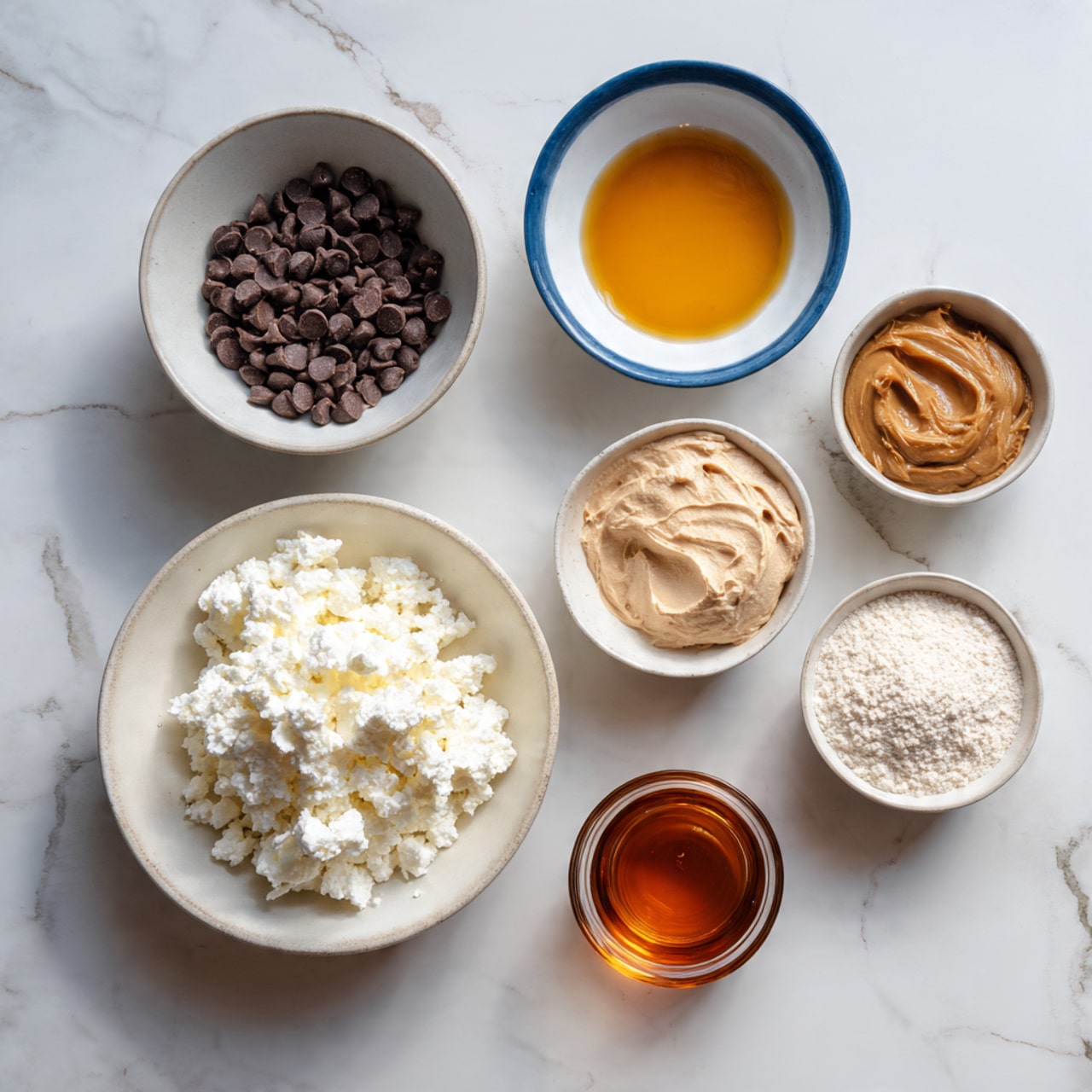 The image shows seven bowls on a white marbled surface, each containing different ingredients. Starting from the top left, there is a white bowl filled with small dark brown chocolate chips. To the right of it is a smaller white bowl with a blue rim holding a golden amber liquid. Below that, there is a white bowl with a creamy light brown powder. Next to it, on the right, is another white bowl with a thick, caramel-colored paste. Below the chocolate chips is a white bowl filled with white cottage cheese with a lumpy texture. To the right of it, a white bowl contains a light beige powdery flour. Below the cottage cheese bowl, there is a small glass cup with a rich amber liquid. The setup is neat, with the ingredients evenly spaced out on the white marbled surface. photo taken with an iphone --ar 4:5 --v 7