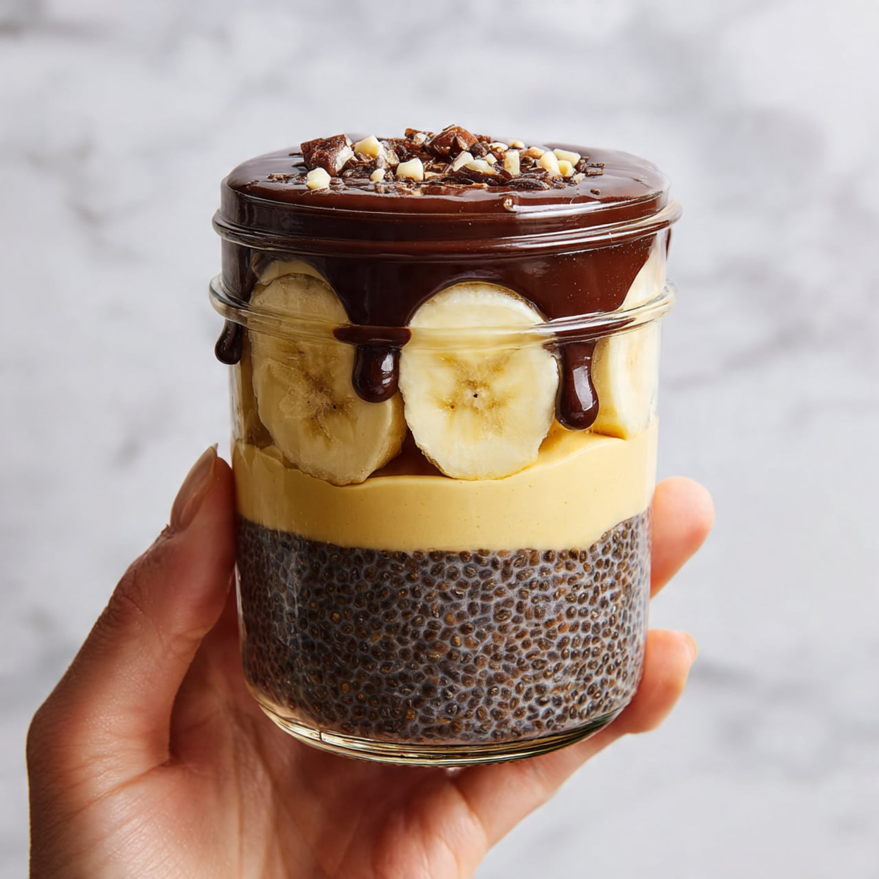 A clear jar held by a woman's hand shows a layered dessert with four distinct layers. The bottom layer is dark brown chia pudding with a grainy texture. Above that is a smooth layer of yellow banana slices, arranged evenly across the jar. The third layer is another thick portion of the dark brown chia pudding, matching the first. The top layer is a creamy beige substance, smooth and filling the jar’s top. On the very top, shiny dark brown chocolate sauce drips slightly over the edge, creating a glossy finish. The jar is set against a white marbled surface. photo taken with an iphone --ar 4:5 --v 7
