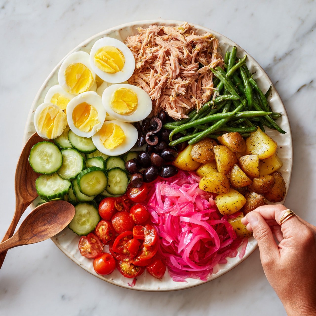 The dish is a large white divided plate filled with six different layers of food. On the top left side, there are sliced boiled eggs with soft yellow yolks, arranged neatly. Next to the eggs on the top right, there is a pile of shredded brown tuna. Below the tuna, on the right side, are small roasted potatoes with a golden-brown color. In the center and slightly to the right, there are green beans mixed with bright pink pickled onions. On the bottom left, there are sliced cucumbers in a light green shade. In the middle left section are dark black olives and red cherry tomatoes, both cut in half. The plate sits on a white marbled surface, and two wooden salad spoons along with a woman’s hand are positioned on the bottom right side of the image. The scene is lit naturally, showing the fresh and colorful textures of the food, photo taken with an iphone --ar 4:5 --v 7