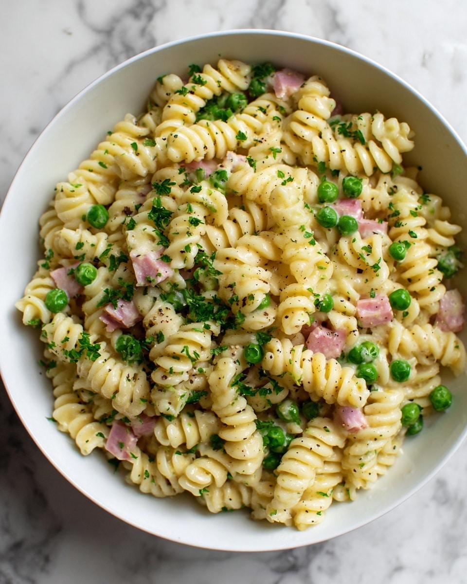 A white bowl filled with creamy pasta made of spiral rotini noodles mixed evenly with small green peas and tiny pink pieces of ham. The pasta is coated in a smooth, light cream sauce with visible specks of black pepper sprinkled on top. Small bright green parsley leaves are scattered throughout, adding a fresh touch. The bowl sits on a white marbled surface. photo taken with an iphone --ar 4:5 --v 7