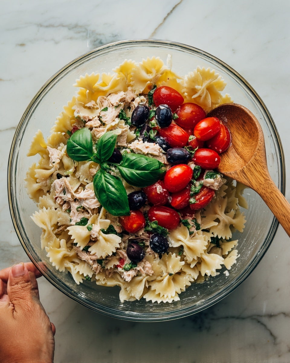 A clear glass bowl filled with three layers of farfalle pasta mixed with chopped black olives, halved bright red cherry tomatoes, chunks of light pink tuna, and fresh green basil leaves on top. The pasta is creamy white with a smooth texture, the tomatoes are firm and shiny, and the olives are dark purple and small. A wooden spoon is stirring the mixture from the right side, held by a woman's hand with a light skin tone, and the bowl is placed on a white marbled surface. photo taken with an iphone --ar 4:5 --v 7