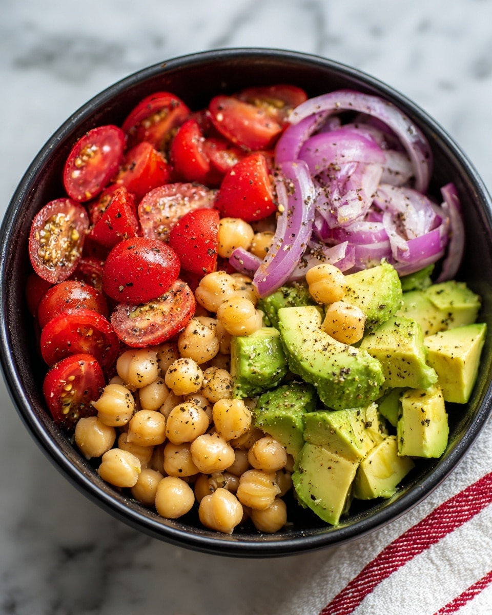 A black bowl on a white marbled surface holds a colorful salad with three main layers. The bottom layer is made of round beige chickpeas, scattered throughout. On top of the chickpeas are bright red, halved cherry tomatoes and medium green avocado chunks with a smooth texture. Thin slices of purple-red onion are mixed in, adding a slight curve and layering effect. The salad is lightly sprinkled with coarse black pepper, enhancing the visual texture. Part of a white and red striped cloth is visible near the bowl, and the scene is bright and clear. photo taken with an iphone --ar 4:5 --v 7