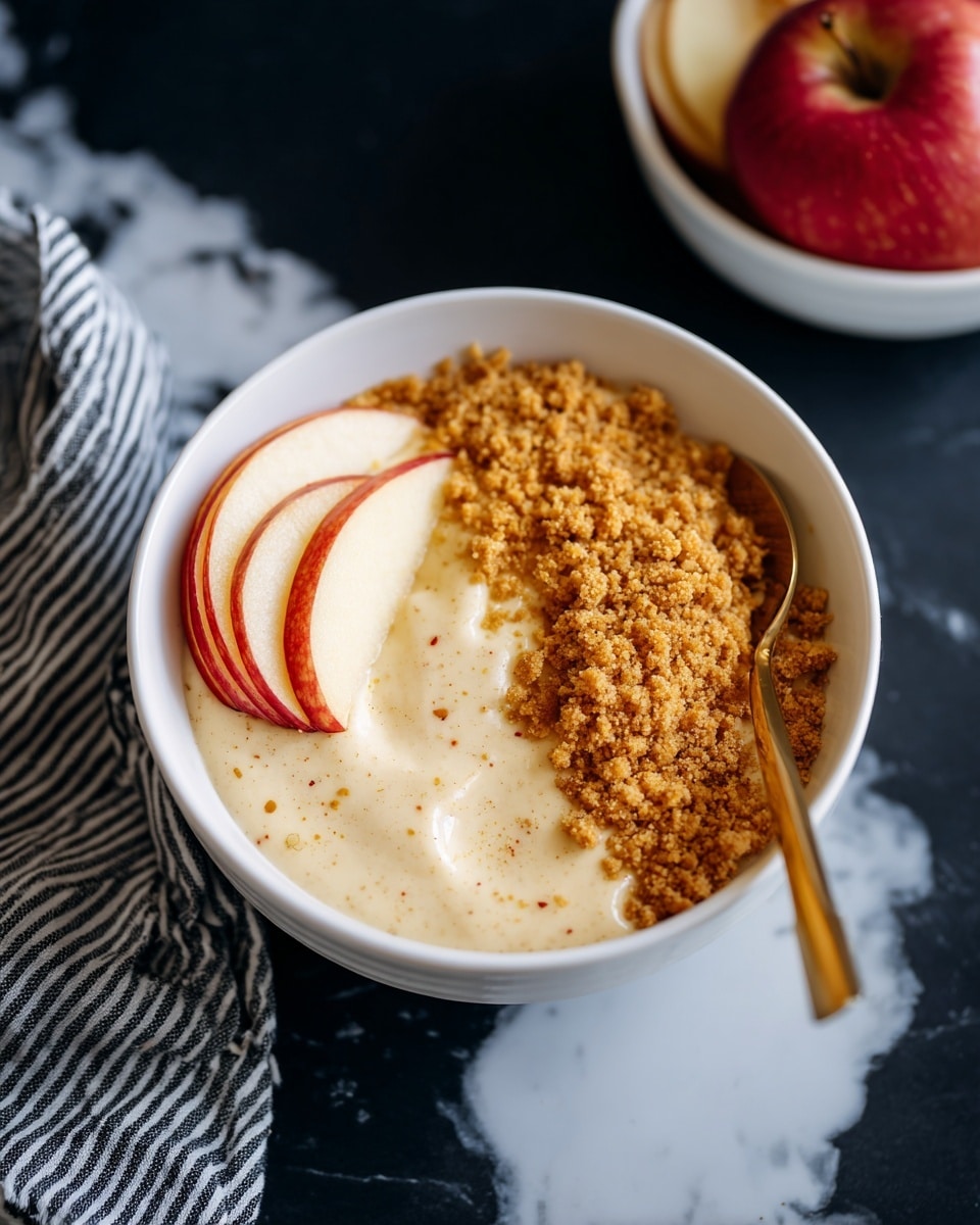 A white bowl shows a creamy mixture in the bottom half with a light beige color and soft, smooth texture. Above the creamy part is a layer of crushed crumbs spread evenly, golden brown in color. On the left side of the bowl, one slice of a fresh apple rests partly inside the bowl, showing its pale yellow and red skin. A gold spoon is placed inside the bowl on the right side, partly submerged in the creamy mix. The bowl sits on a dark surface with a white marbled texture, with a red apple and another white bowl of sliced apples nearby. A striped black and white cloth is on the left side. Photo taken with an iphone --ar 4:5 --v 7