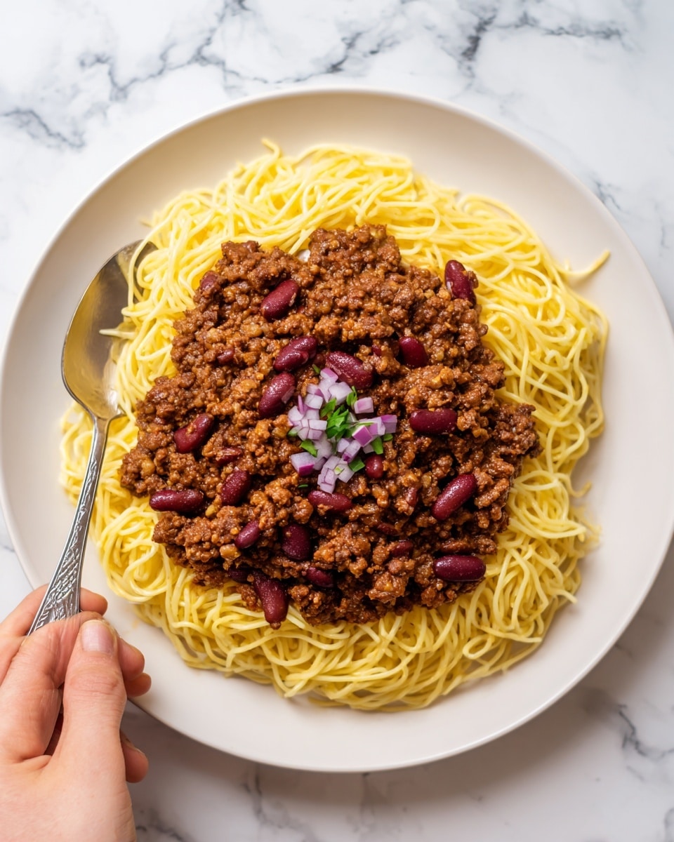 The image shows a white plate filled with thin yellow spaghetti noodles as the bottom layer. On top of the noodles, there is a thick layer of dark brown ground meat sauce covering most of the pasta. Scattered over the sauce are small red kidney beans and light tan round crackers. There are also a few pieces of chopped purple onions placed on top in a small cluster. A woman's hand is holding a spoon near the edge of the plate. The plate sits on a white marbled textured surface. photo taken with an iphone --ar 4:5 --v 7