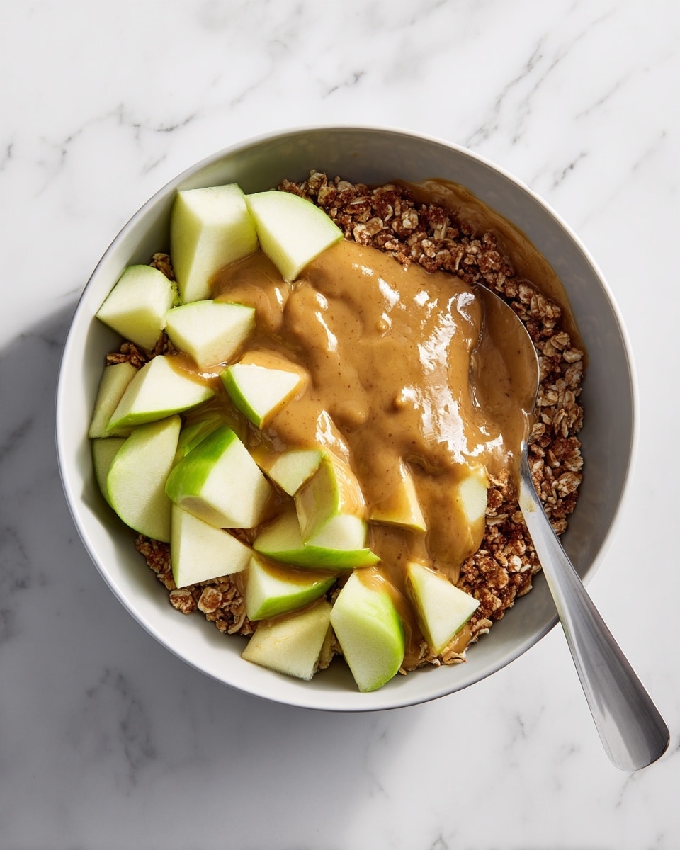 A white bowl filled with three main layers. The bottom layer is a bed of soft brown oats. On top of the oats are several chunks of green apple with shiny skins and pale inside. The final layer is a smooth, creamy brown sauce that covers the apples and oats, with some sauce dripping slightly over the sides. The bowl is placed on a white marbled surface, and a spoon is partially visible on the left side inside the bowl. photo taken with an iphone --ar 4:5 --v 7