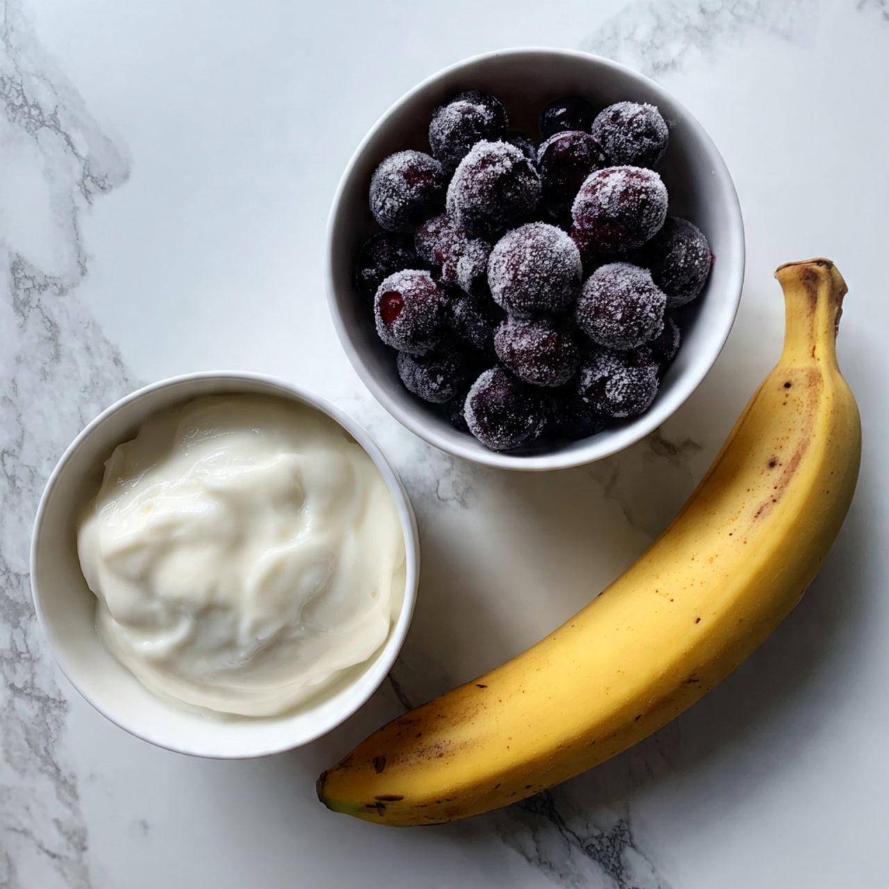 The image shows three items arranged on a white marbled surface: a white bowl filled with smooth, creamy white yogurt on the left, a white bowl filled with dark purple frozen blueberries covered lightly in frost at the top right, and a whole ripe yellow banana with a few brown spots placed diagonally on the lower right side. Photo taken with an iphone --ar 4:5 --v 7