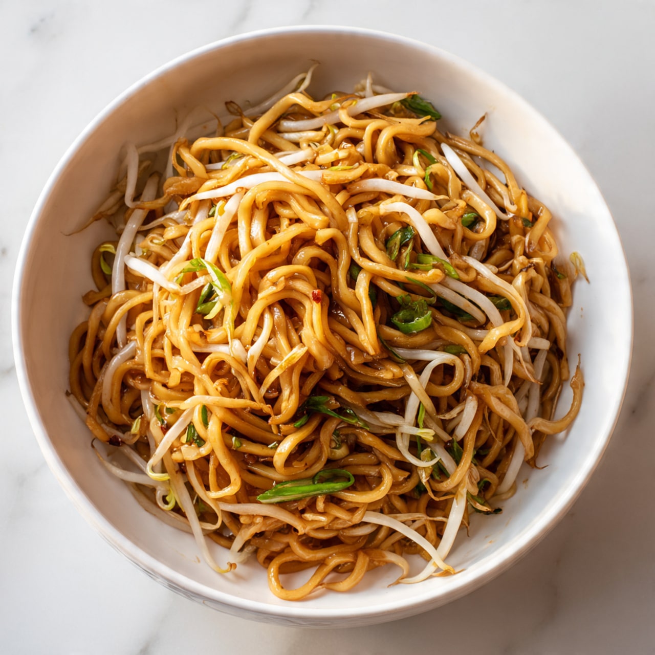 This image shows a white bowl filled with a single serving of stir-fried noodles. The noodles are thick and twisted together, with a rich light brown color from the sauce coating them. Mixed in are thin, white bean sprouts and small pieces of green onion, adding light green and off-white touches throughout the noodles. The bowl is placed on a white marbled surface, giving a clean and bright look to the setting. photo taken with an iphone --ar 4:5 --v 7
