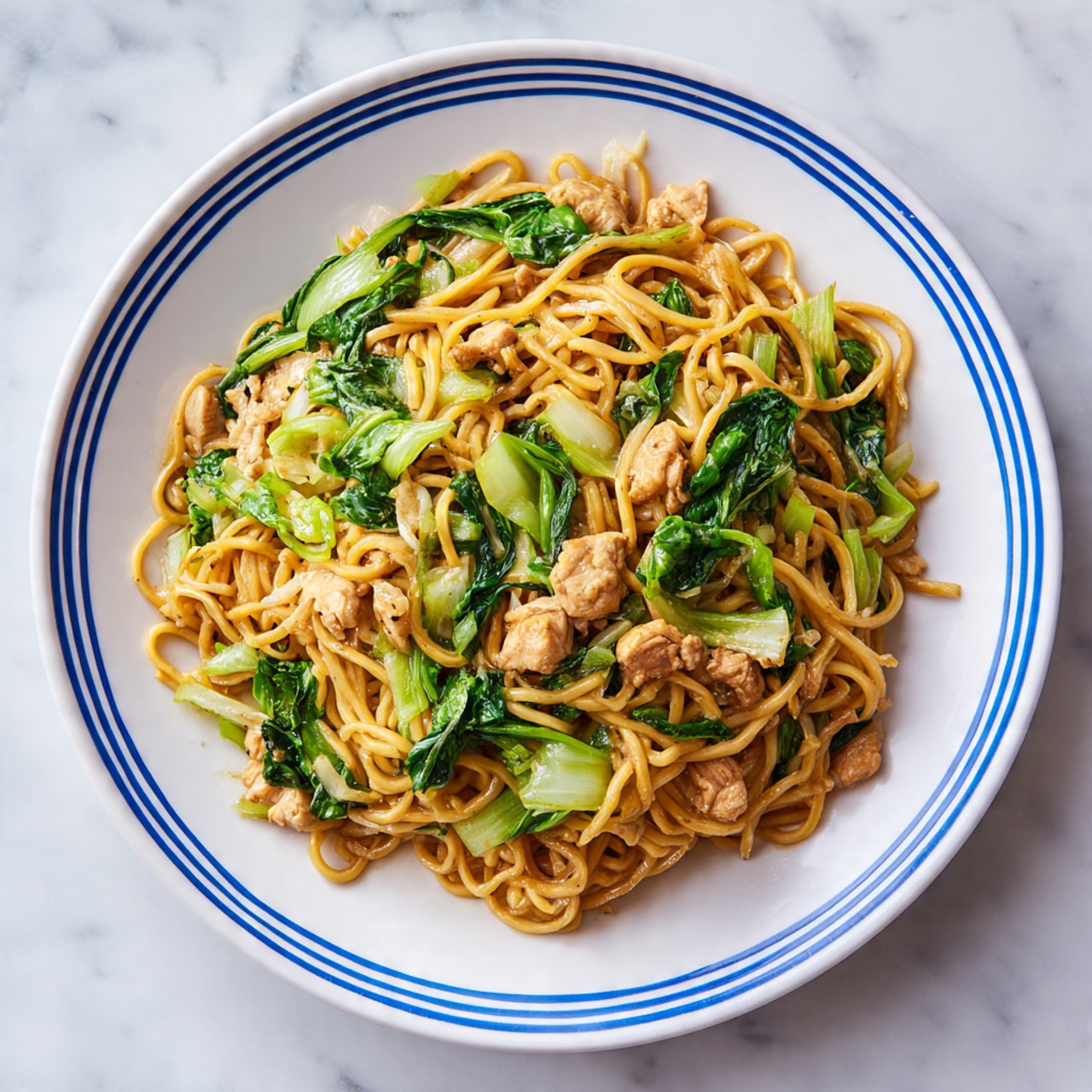 A white plate with blue stripes on the rim holds a large pile of stir-fried noodles mixed with small pieces of light brown chicken and bright green leafy vegetables. The noodles look soft and slightly shiny, with a golden-brown color from the sauce. The chicken pieces are scattered evenly throughout, and the green vegetables add a fresh contrast. The plate sits on a white marbled surface. photo taken with an iphone --ar 4:5 --v 7