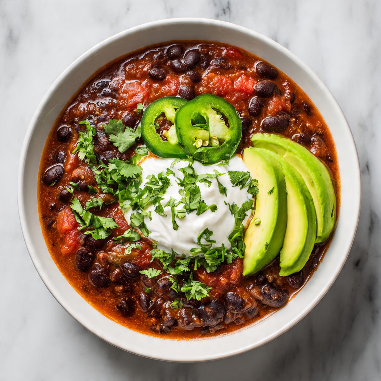 A white bowl filled with a thick soup made of black beans and red tomatoes, topped with a dollop of white sour cream in the center. There are three bright green slices of avocado neatly placed on one side of the bowl. On top of the sour cream, there are two round green slices of jalapeño and small pieces of fresh green cilantro sprinkled around. The bowl is set on a white marbled surface. Photo taken with an iphone --ar 4:5 --v 7