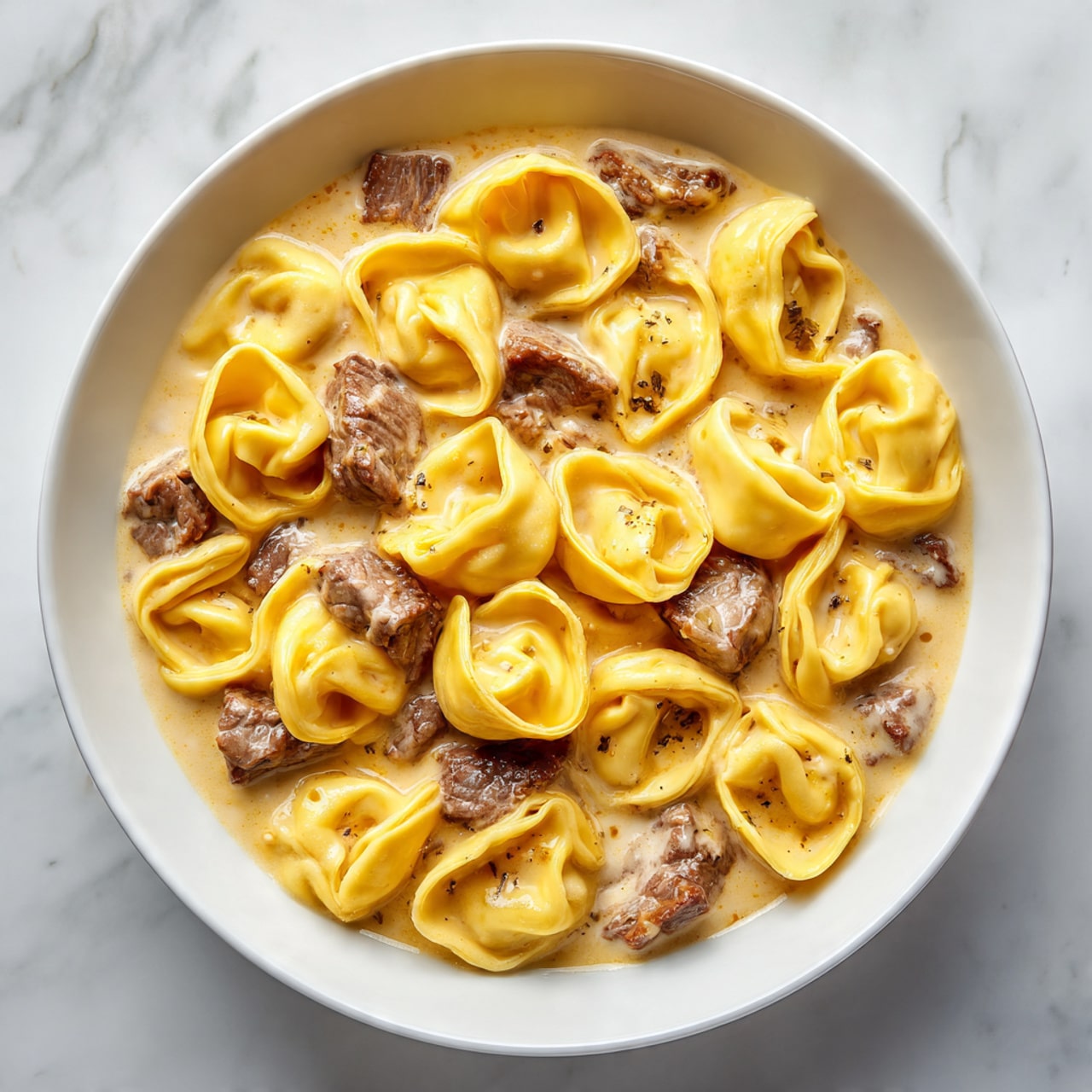 The image shows a white bowl filled with creamy sauce holding many yellow tortellini pasta and pieces of brown beef. The tortellini are plump and smooth, arranged evenly throughout the creamy sauce that has a slight shine. The beef is scattered between the pasta, adding texture with its irregular shape and slightly browned edges. The creamy sauce looks thick and coats both pasta and beef well. The background surface is white marbled texture. Photo taken with an iphone --ar 4:5 --v 7