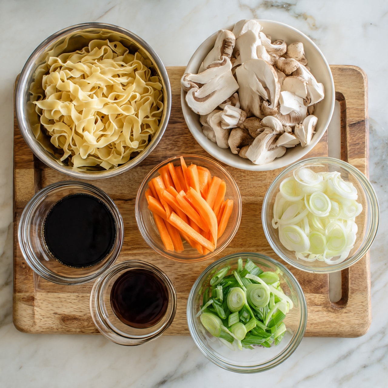The image shows six containers with different food items placed on a light brown wooden board over a white marbled surface. In the top left, there is a metal bowl holding pale yellow cooked noodles with a smooth texture. To the top right, a white bowl is filled with white and light brown sliced mushrooms showing soft, rounded edges. In the center, a small clear glass bowl contains thin, bright orange carrot sticks. Below the carrots, two more small glass bowls hold light green sliced green onions on the right and white onion slices on the left, both with a fresh and crisp look. Finally, to the left bottom corner, there is a small clear glass bowl with dark soy sauce, smooth and shiny on the surface. Photo taken with an iphone --ar 4:5 --v 7