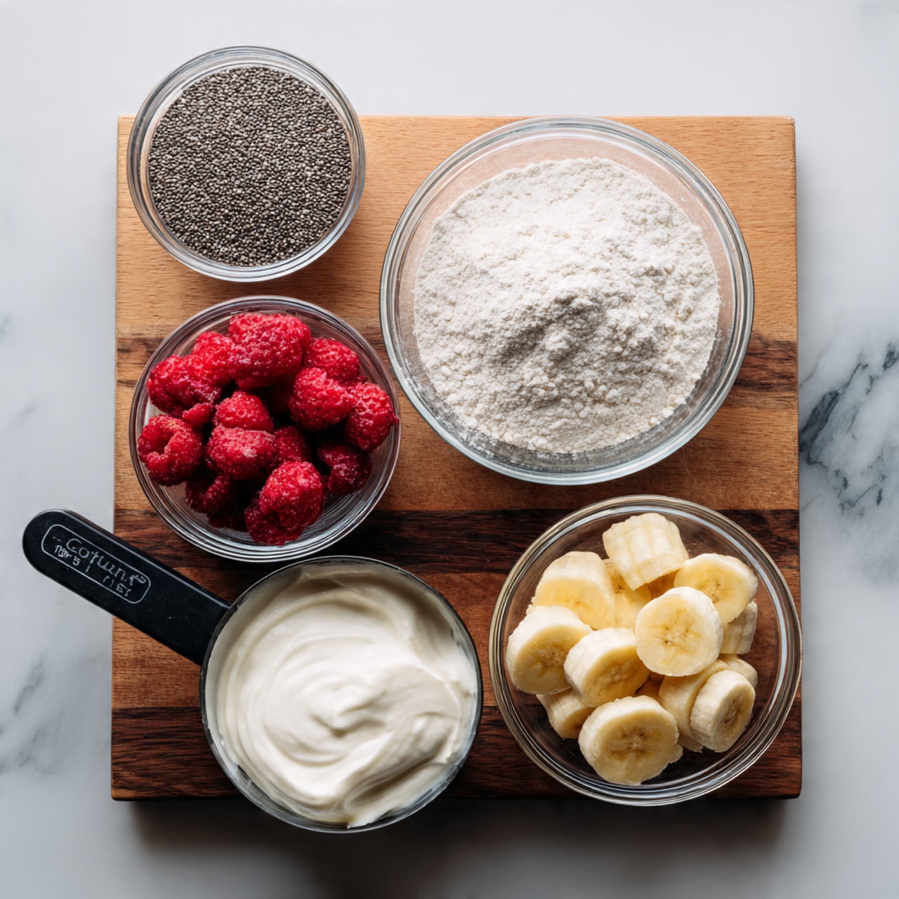 A top view of six glass bowls and a metal measuring cup arranged on a small wooden board on a white marbled surface. The top left bowl contains small black chia seeds with a rough texture. Below it, a larger bowl is filled with fine white flour, smooth and powdery. To the right of the flour, a metal measuring cup holds fresh bright red raspberries with a soft, bumpy surface. Below the measuring cup, a medium bowl is filled with frozen red strawberries that have icy textures. At the bottom right, a smaller glass bowl contains pale yellow banana slices with a soft texture. Finally, at the bottom left, a black measuring cup is heaped with white creamy yogurt that looks thick and smooth. photo taken with an iphone --ar 4:5 --v 7