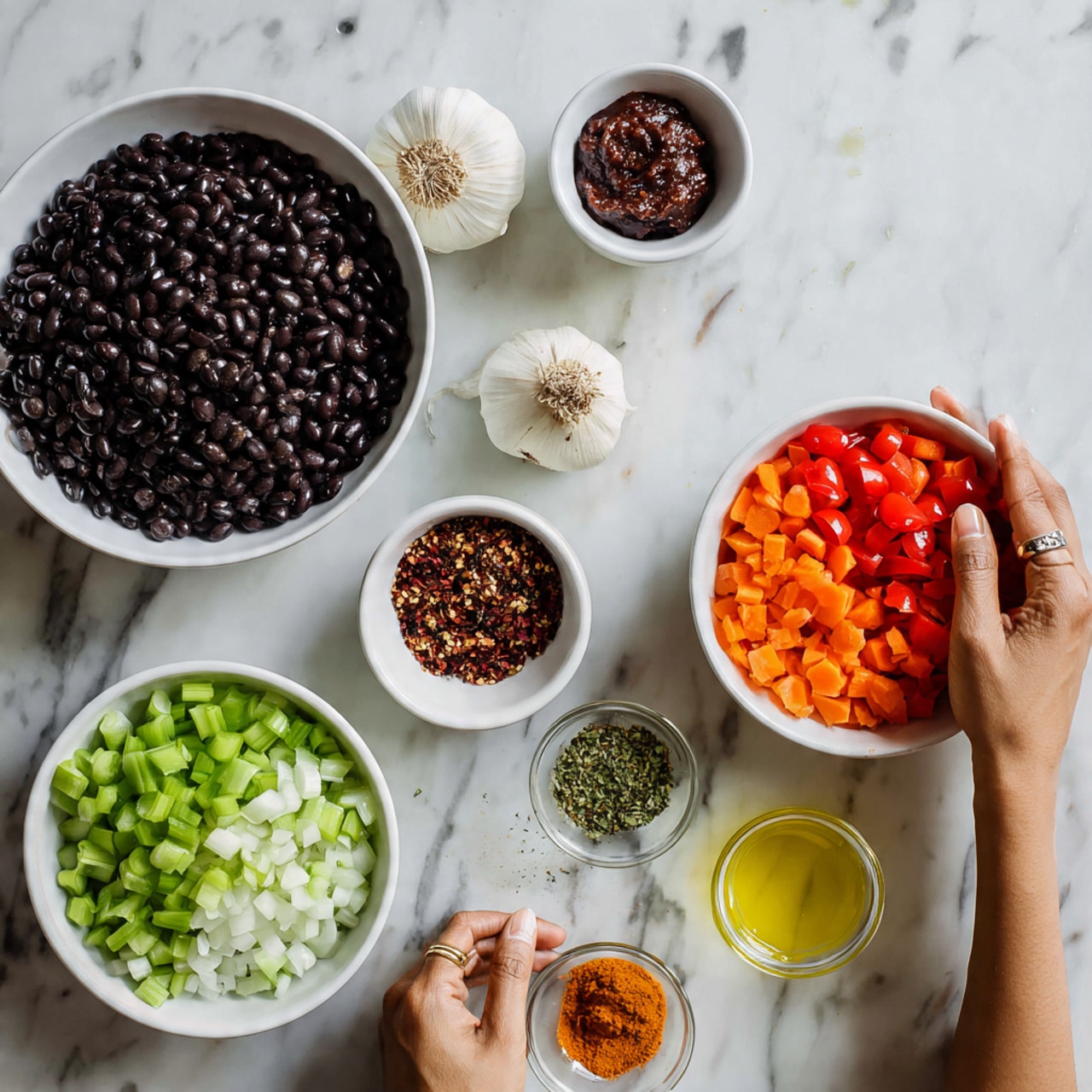 The image shows several white bowls on a white marbled surface. One large white bowl is filled with black beans. Another large white bowl is divided into sections holding chopped green celery, red tomatoes, orange carrots, and white onions. A small white bowl holds spices of different colors including green, red, and brown. There is a small clear bowl with a dark red paste and a clear glass container with yellow liquid. A whole garlic bulb is placed near the bowls. A woman's hand is gently holding one of the bowls. photo taken with an iphone --ar 4:5 --v 7