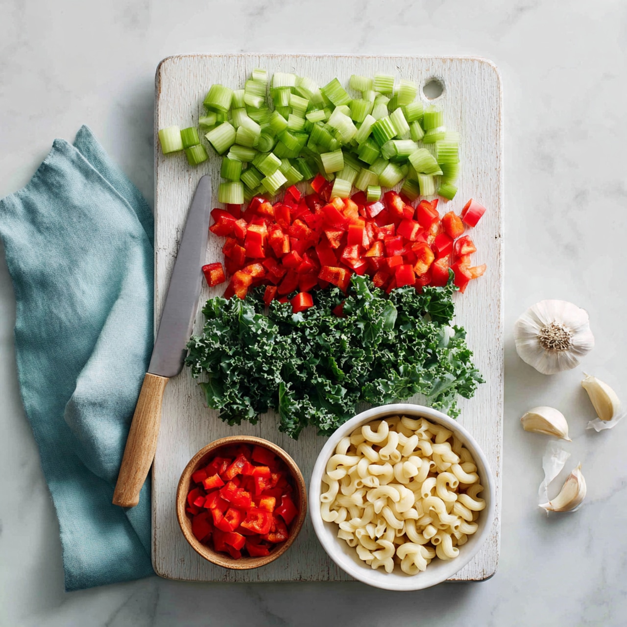 The image shows a white wooden board on a white marbled surface with neatly chopped vegetables arranged in rows: green celery at the top, chopped red bell peppers below, then chopped kale, and more red bell peppers in a small wooden bowl at the bottom. To the front right of the board is a white bowl filled with uncooked macaroni pasta. A knife with a wooden handle lies on the board beside the vegetables. A pale blue cloth is folded on the left side of the board, and two cloves of garlic rest on the marbled surface to the right. The scene is bright and clean, showing fresh ingredients ready for cooking, photo taken with an iphone --ar 4:5 --v 7