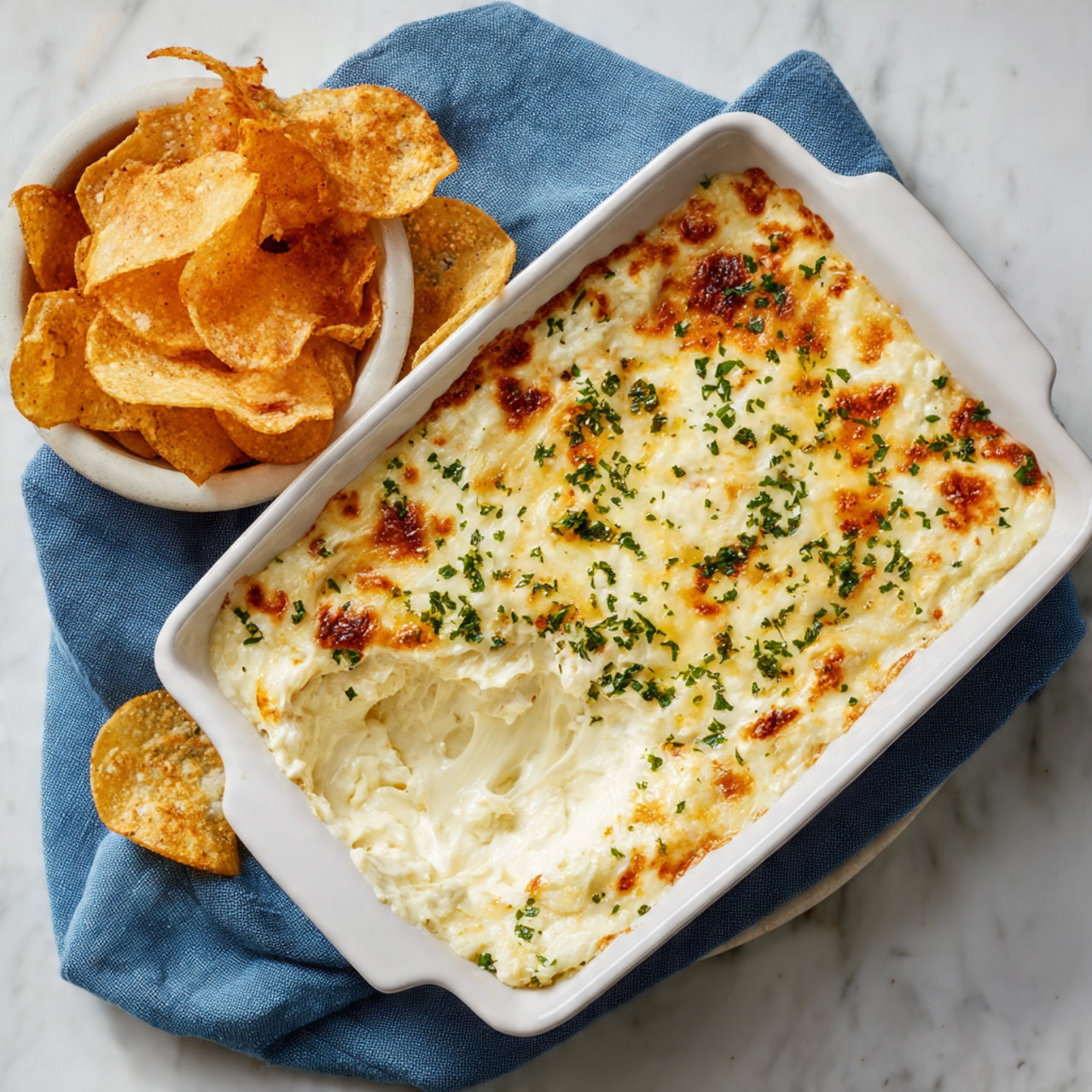 A white rectangular dish filled with a creamy dip topped with melted golden brown cheese and sprinkled with chopped green herbs. On the side, there is a white bowl filled with crispy fried chips. The dip has a smooth texture under the cheese layer with some areas showing a soft, cheesy pull where a scoop is taken out. The whole setting is placed on a white marbled surface with a blue cloth underneath the dish. Photo taken with an iphone --ar 4:5 --v 7