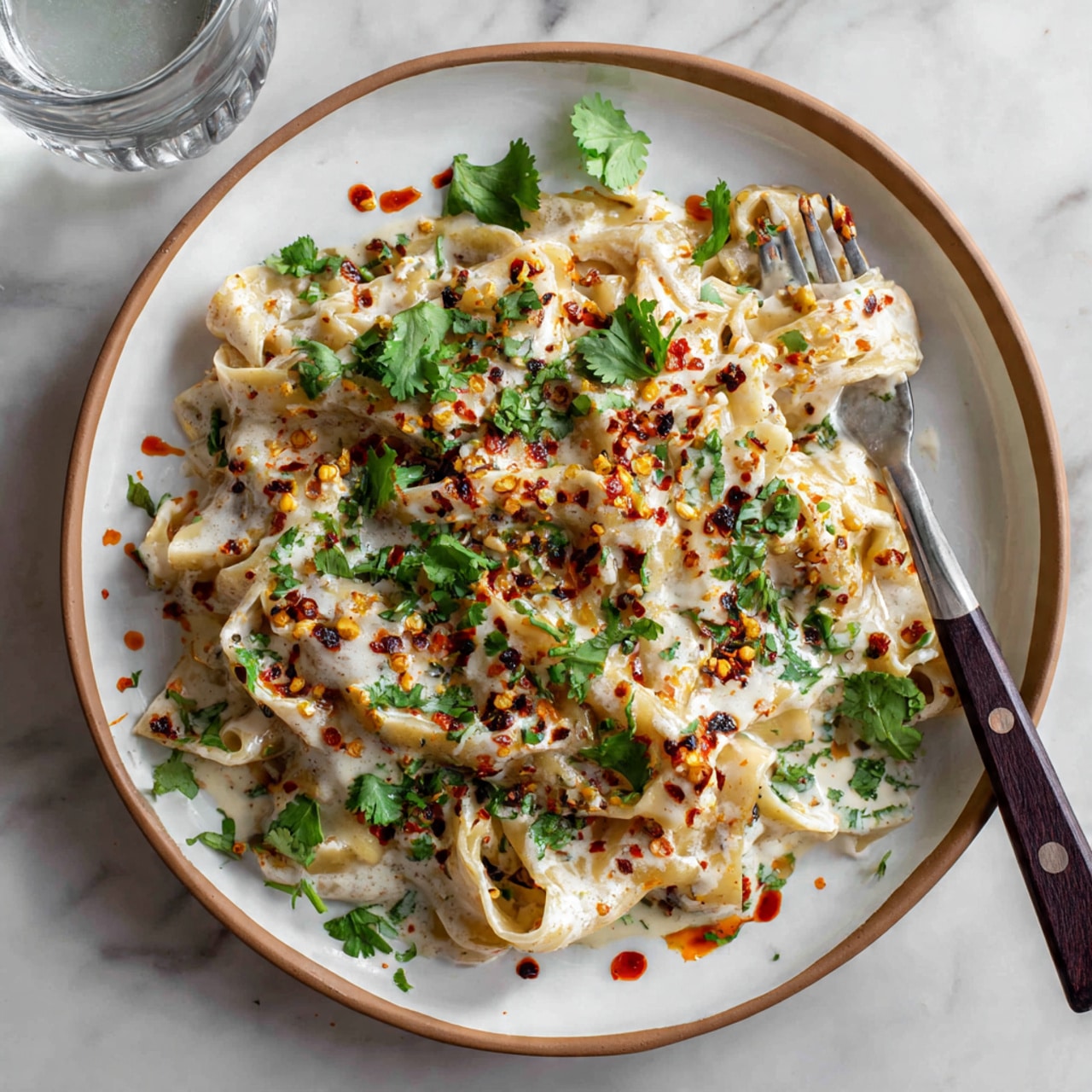 This image shows a white plate with a light brown rim filled with flat noodles covered in a creamy white sauce. The noodles are topped with green chopped herbs and scattered fresh green cilantro leaves. There are splashes of dark red chili oil with chili flakes, giving spots of red and orange around the noodles. A fork with a dark brown handle rests on the plate, holding some noodles. The background is a white marbled surface with a clear glass of water nearby. Photo taken with an iphone --ar 4:5 --v 7