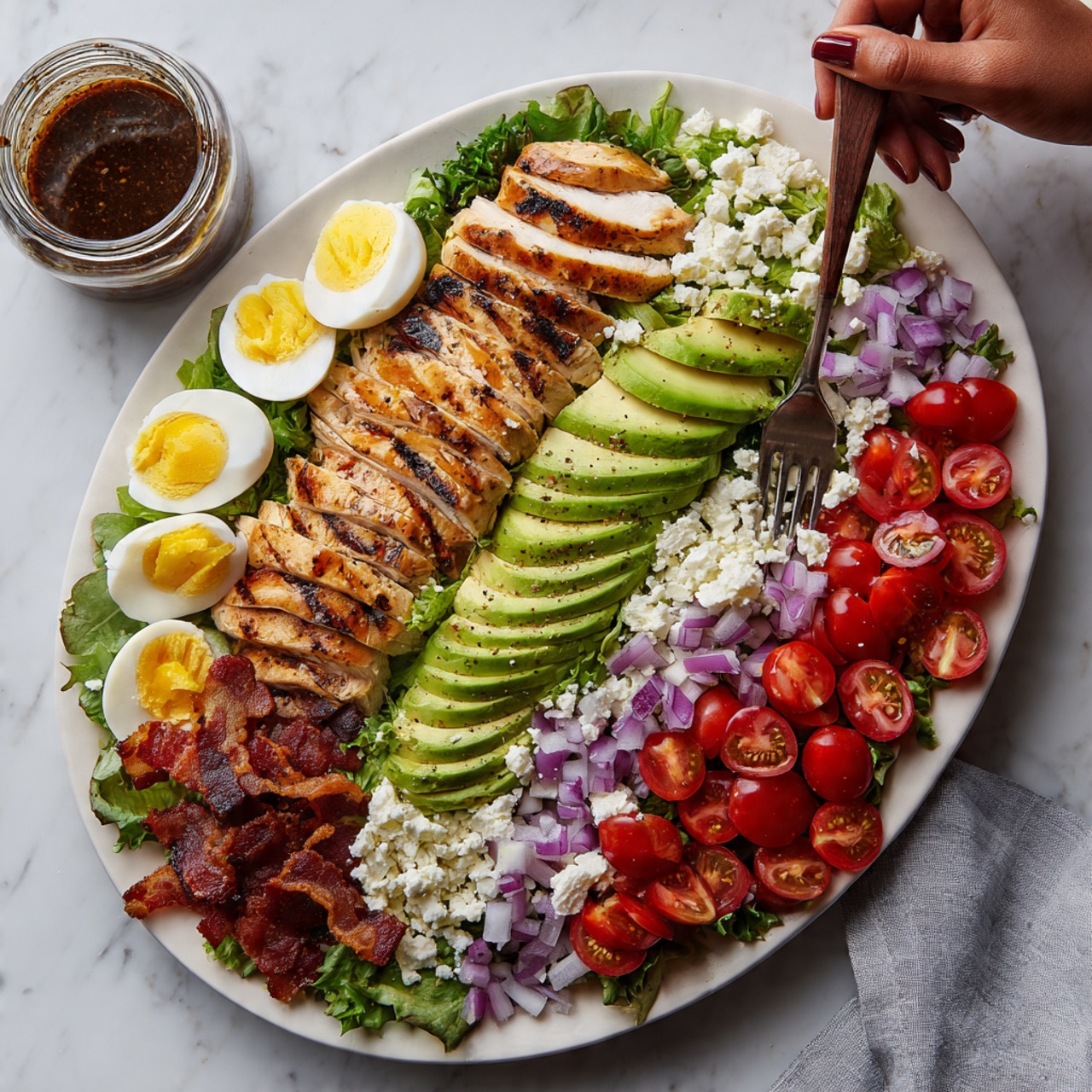 The dish shows a large white oval plate on a white marbled surface, with neatly arranged layers of food. On the left and right sides are rows of light golden grilled chicken slices with a smooth texture. Next to the chicken are bright green avocado slices, fanned out evenly. Cherry tomatoes, halved and red, are scattered in small groups along the edges. In the center is a layer of soft-looking crumbled white cheese. On one side of the cheese are finely chopped purple onions and green lettuce leaves, while crispy, dark brown bacon pieces line the outer edges. At the bottom left corner, there are wedges of hard-boiled eggs with white and bright yellow yolks. A jar with dark dressing and a wooden spoon are nearby, with a woman's hand holding a fork over the plate. Photo taken with an iphone --ar 4:5 --v 7