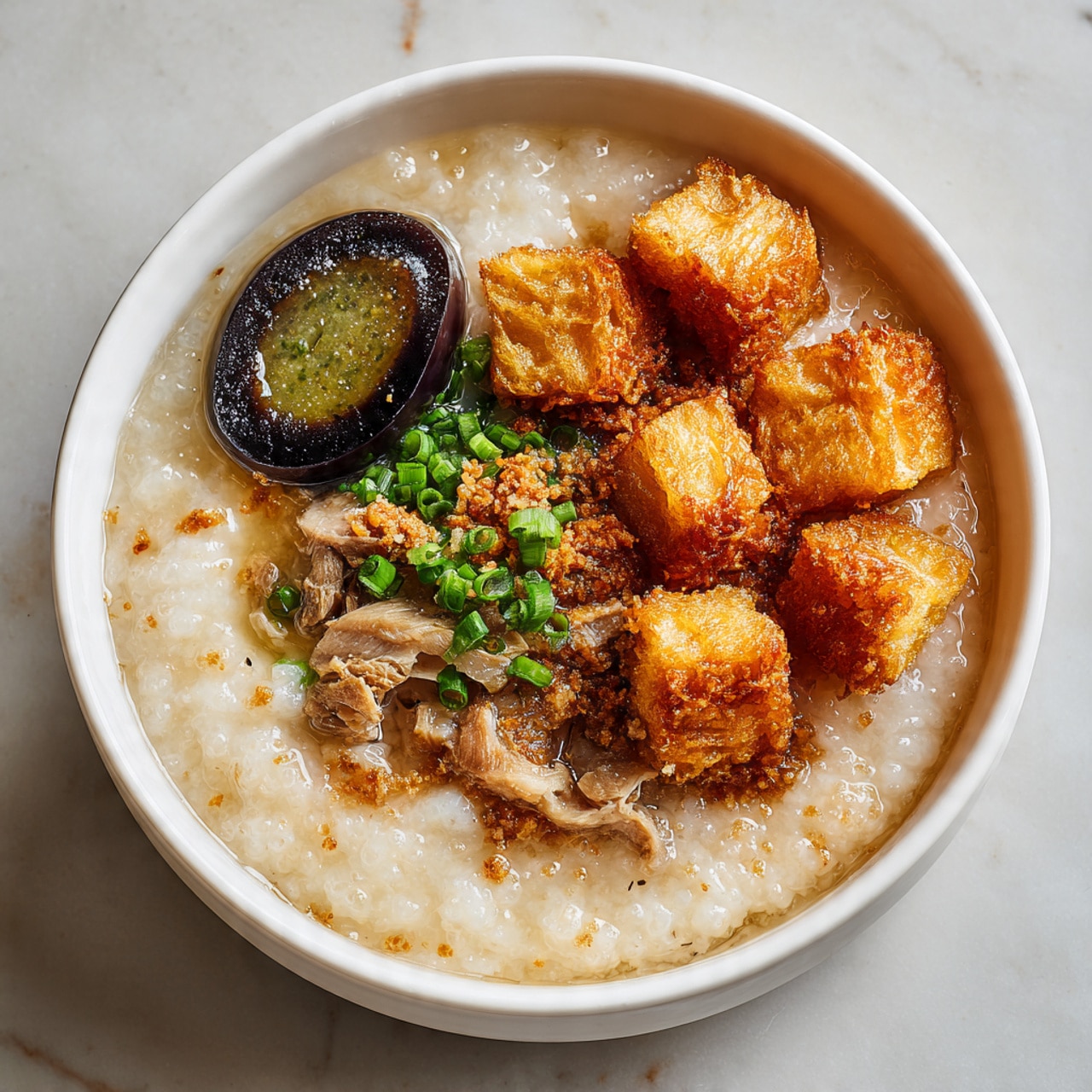 A white bowl filled with thick, creamy rice porridge that has a soft, smooth texture in the base layer. On top, there are several small pieces of golden, crispy fried dough blocks clustered on one side. To the left, a dark, glossy century egg with a marbled green and black yolk is placed partially soaked in the porridge. Scattered around are small pieces of tender meat mixed into the porridge and a sprinkle of fresh, bright green chopped spring onions adds color and freshness on the right side. The bowl sits on a white marbled surface with soft lighting. Photo taken with an iphone --ar 4:5 --v 7