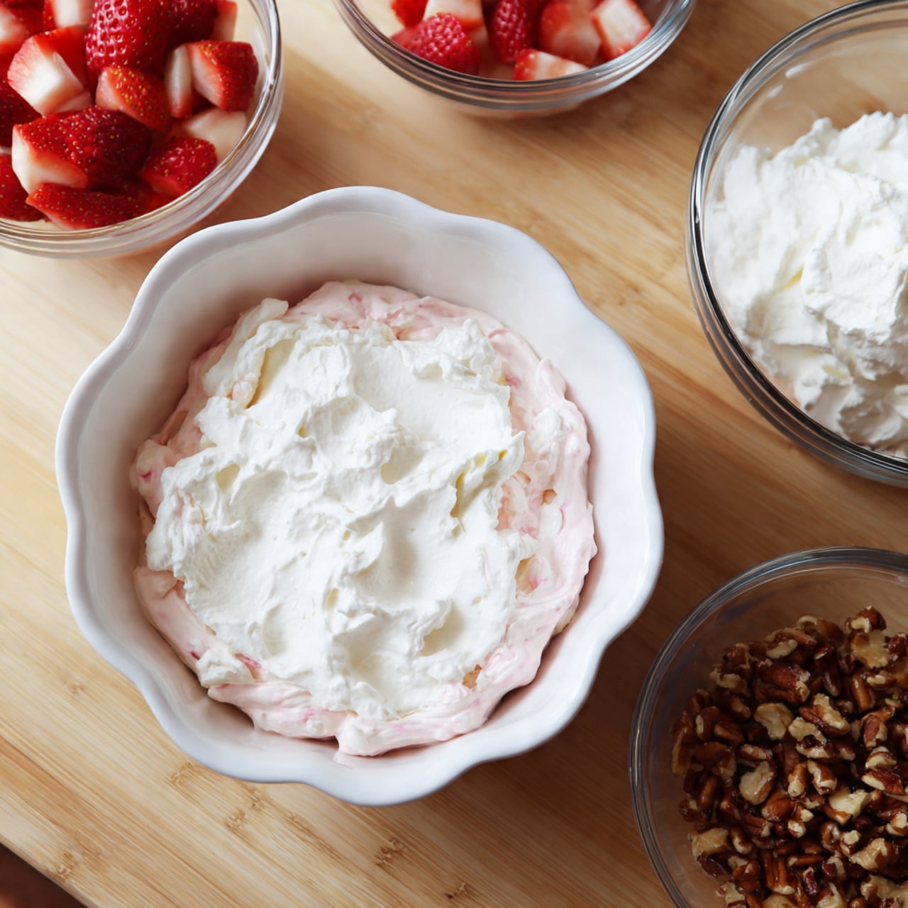 A close-up image shows a dessert preparation in a white scalloped bowl with pink and white creamy mixture topped with fluffy white cream, next to three small clear bowls on a wooden surface. One bowl holds more whipped cream, another contains fresh red and white strawberry pieces, and the last has small brown nut pieces. The scene is bright and simple, focusing on the colorful layers of the dessert and fresh ingredients. Photo taken with an iphone --ar 4:5 --v 7