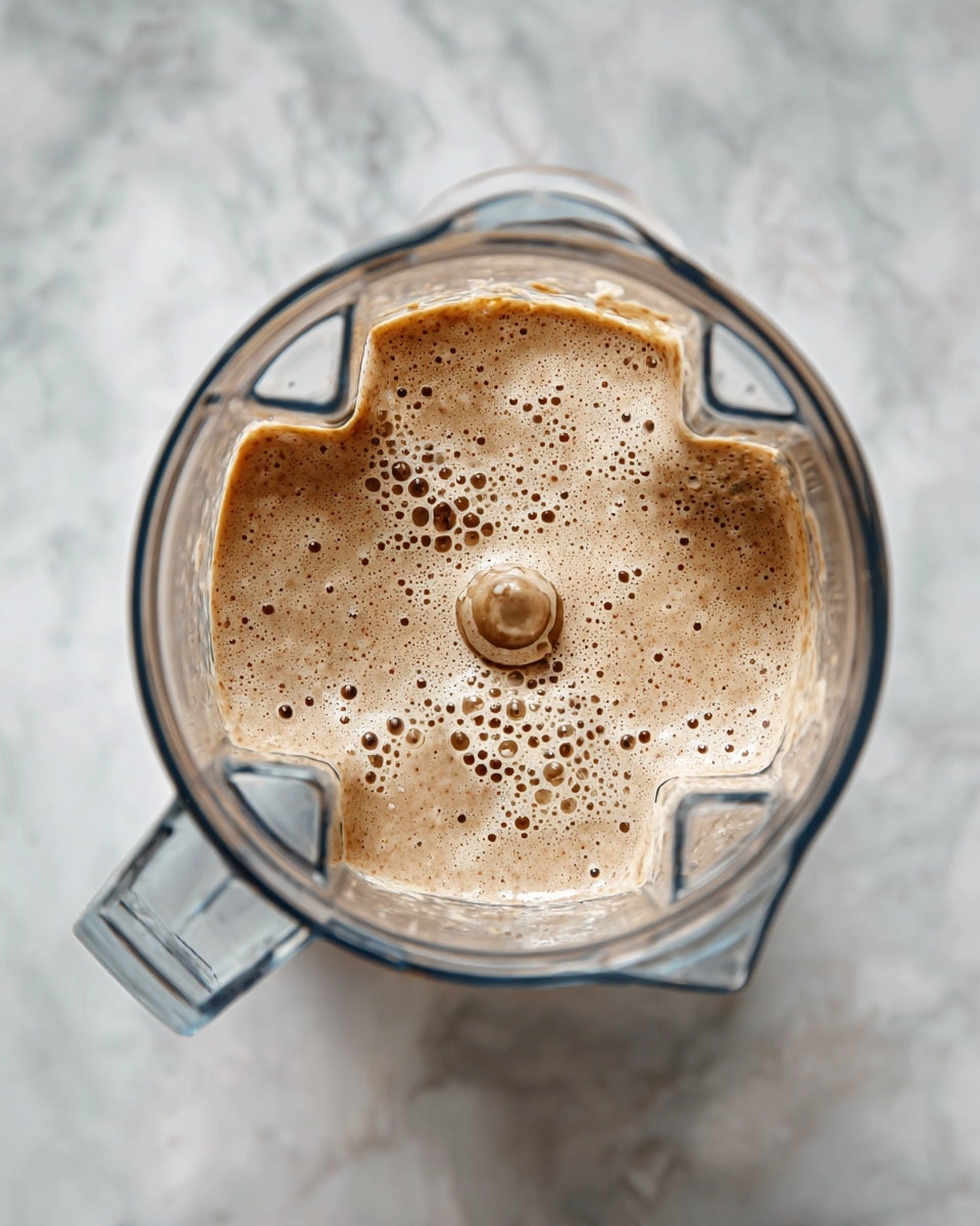 A clear blender container on a white marbled surface filled with a light brown creamy mixture with small darker specks. The mixture's texture is smooth with several bubbles visible on the top, showing it has just been blended. The blender's sharp blades can be seen beneath the liquid inside the container. Photo taken with an iphone --ar 4:5 --v 7