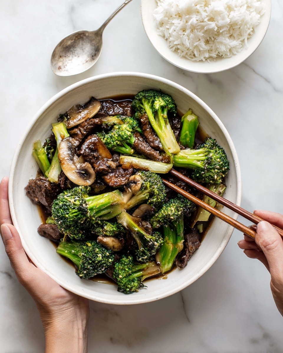 A bowl filled with a stir-fry dish showing layers of green broccoli florets, brown pieces of beef, and light brown sliced mushrooms, all coated in a shiny, dark brown sauce. The bowl is white and round, sitting on a white marbled surface. Nearby, there is a small white bowl filled with white rice and a spoon beside it. A woman's hand is holding a pair of chopsticks above the stir-fry bowl. photo taken with an iphone --ar 4:5 --v 7