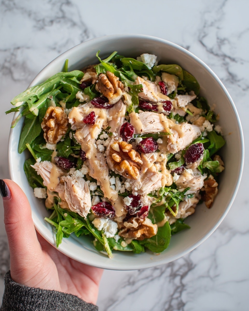 The image shows a white bowl filled with a fresh salad that has several layers. The base layer is dark green leafy arugula. On top of that are chunks of light pink chicken pieces scattered evenly. There are also small white crumbles of cheese sprinkled all over. Bright red dried cranberries are spread across the salad, adding pops of color. Golden brown walnut halves are scattered around, giving texture. The salad is lightly drizzled with a creamy, light brown dressing. A woman's hand is holding the bowl from the side. The background is a white marbled surface. Photo taken with an iphone --ar 4:5 --v 7