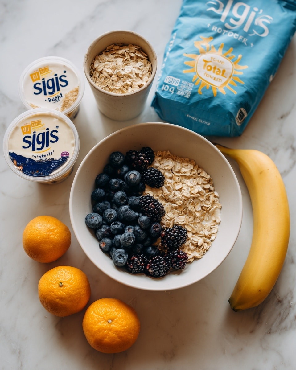 The image shows a white bowl filled with dark blueberries and blackberries placed on a white marbled surface. Surrounding the bowl are a yellow banana laying at the bottom right and a cup filled with oats positioned above the bowl. Next to the oats is a bag of pale blue cereal flakes standing upright. In front of the bowl, there are two small containers of yogurt, one labeled “siggi’s” and the other with