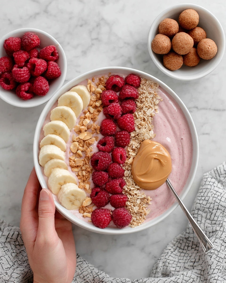 The image shows a white bowl filled with several layers arranged neatly. At the base, there is a smooth pink creamy layer, topped with a row of round banana slices on the left side. To the right of the bananas, there are bright red raspberries piled, with a sprinkle of small light brown granola bits scattered over the middle section. Near the granola, a dollop of light brown peanut butter adds texture and color contrast. Surrounding the main bowl are smaller white bowls, one with more banana slices, another filled with more red raspberries, and a third holding evenly sized round brown balls. The whole setup rests on a white marbled surface, with a gray and white patterned cloth partly visible at the bottom right. A woman’s hand is gently holding a spoon inside the main bowl. Photo taken with an iphone --ar 4:5 --v 7