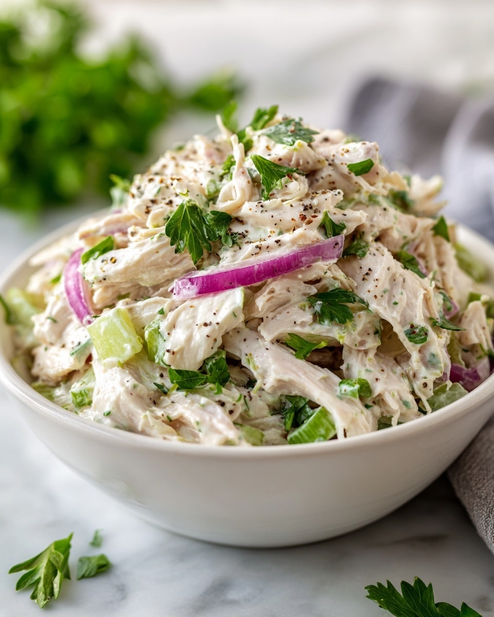 A close-up view of a creamy chicken salad in a white bowl, showing layers of shredded white chicken mixed with light green celery pieces, thin slices of purple-red onion, and a white creamy dressing. The salad is sprinkled with small leaves of fresh green parsley and some cracked black pepper on top, giving texture and color contrast. The bowl is placed on a white marbled surface with some blurred parsley sprigs in the background. photo taken with an iphone --ar 4:5 --v 7